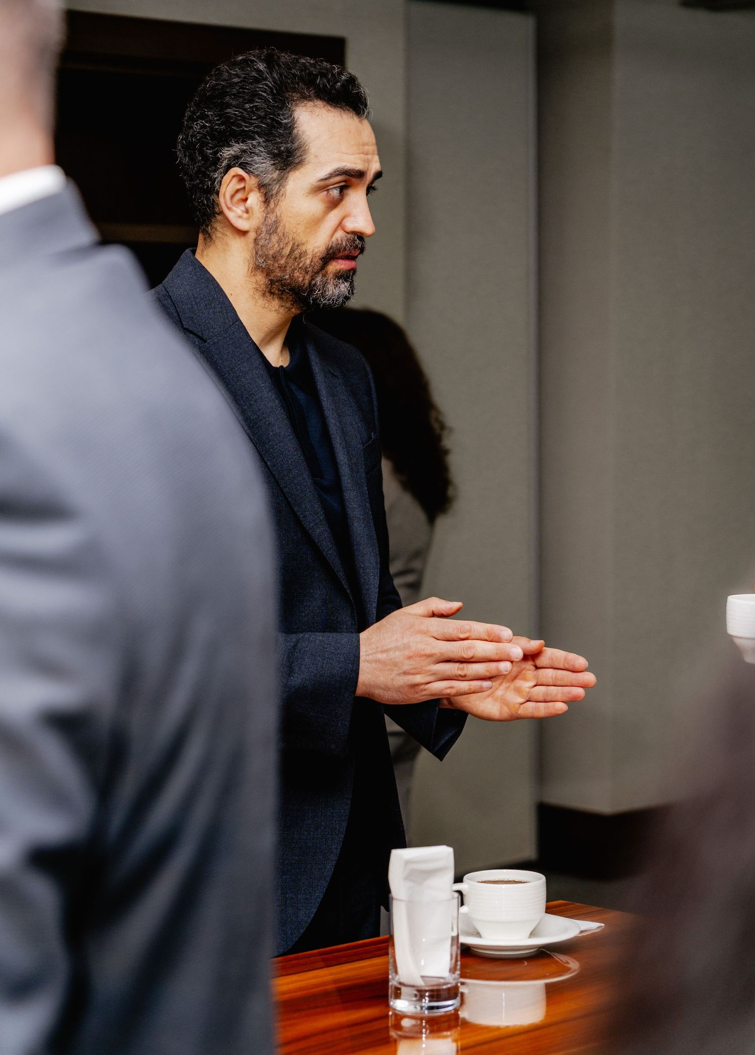 Professional man in dark blazer gesturing while speaking at a business meeting with coffee cups on table.