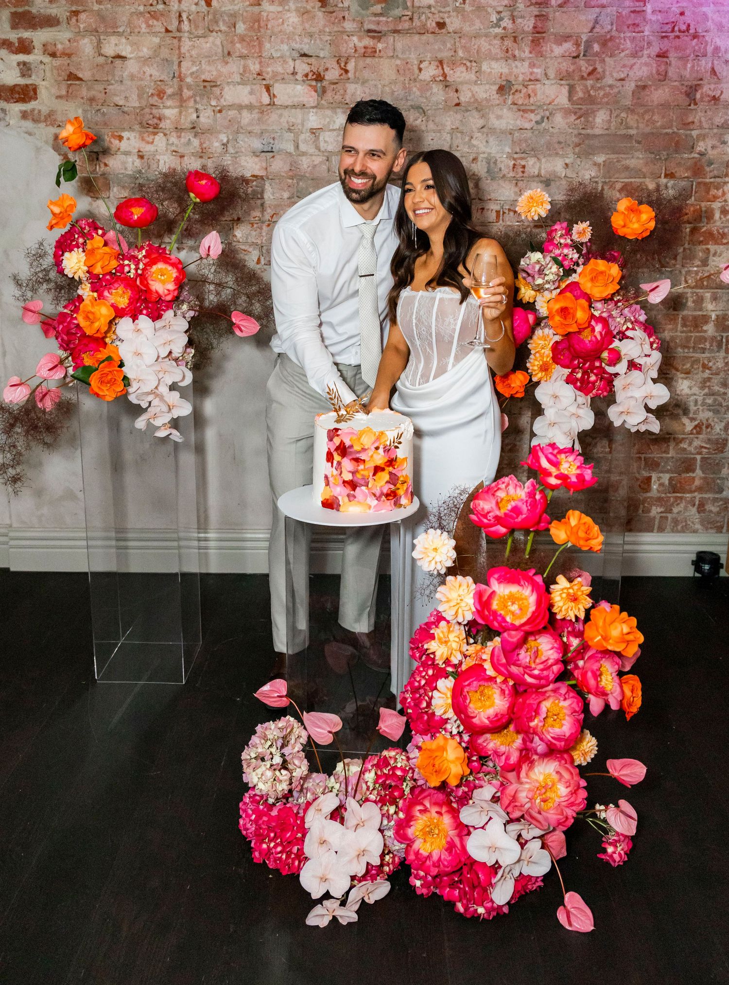 A couple shares a joyful moment against a brick wall decorated with vibrant pink and orange floral arrangements.