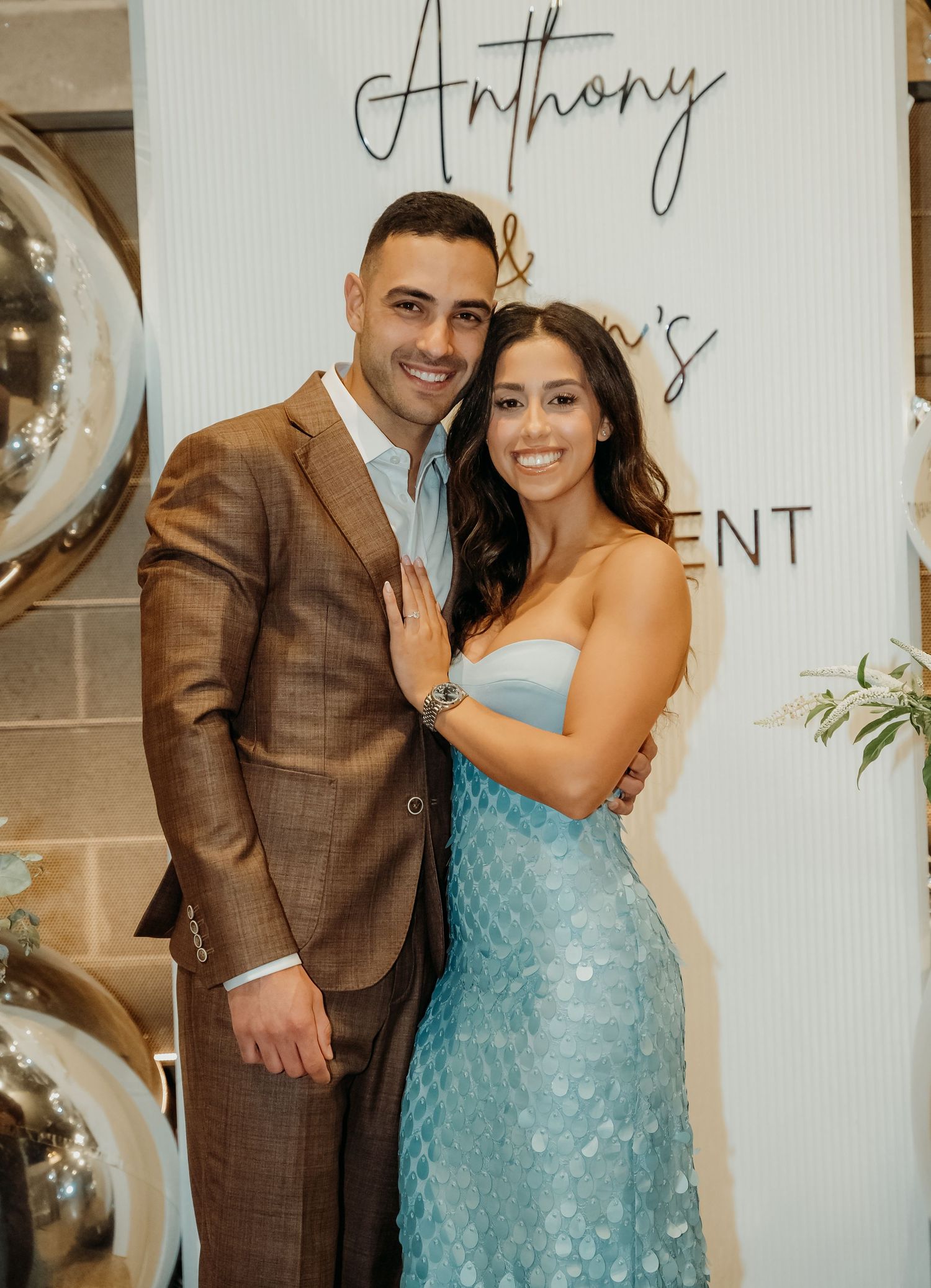 A couple wearing a brown suit and sparkly blue dress embrace joyfully at a wedding celebration.
