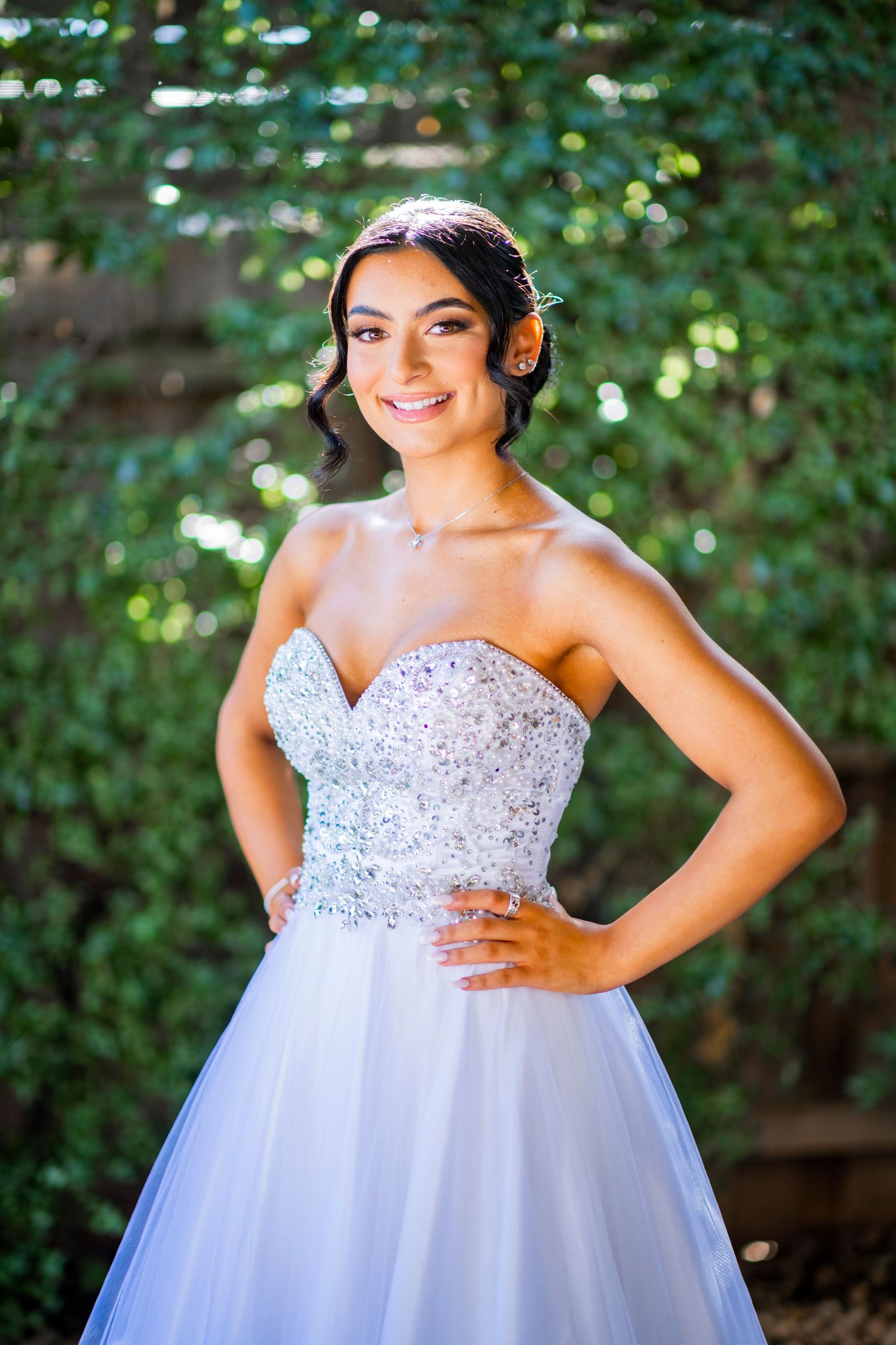 A bride poses in a stunning white ball gown with crystal embellishments and sweetheart neckline against lush greenery.