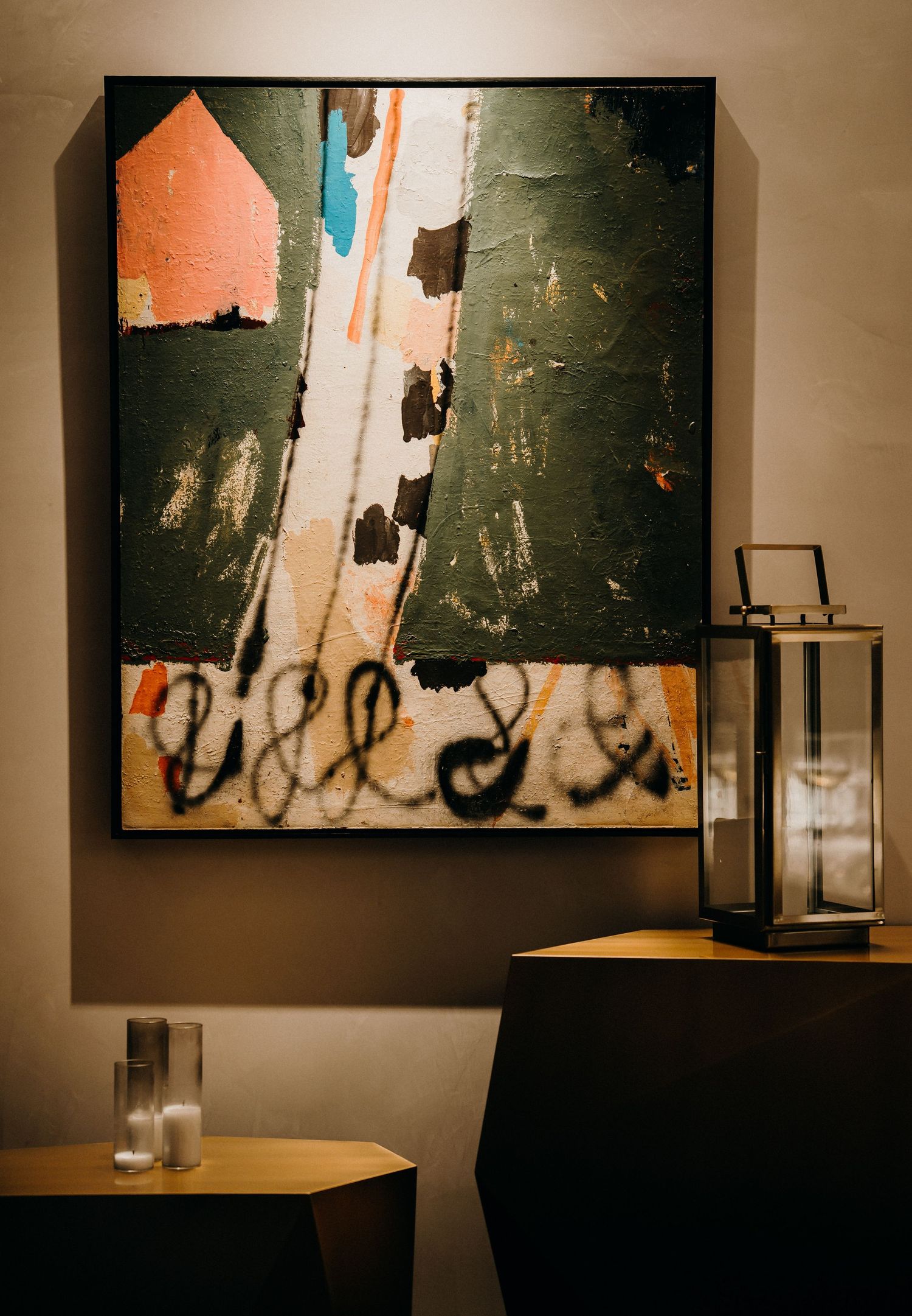 Modern abstract artwork with orange and green tones hangs on wall beside decorative lantern and small table in dim lighting.
