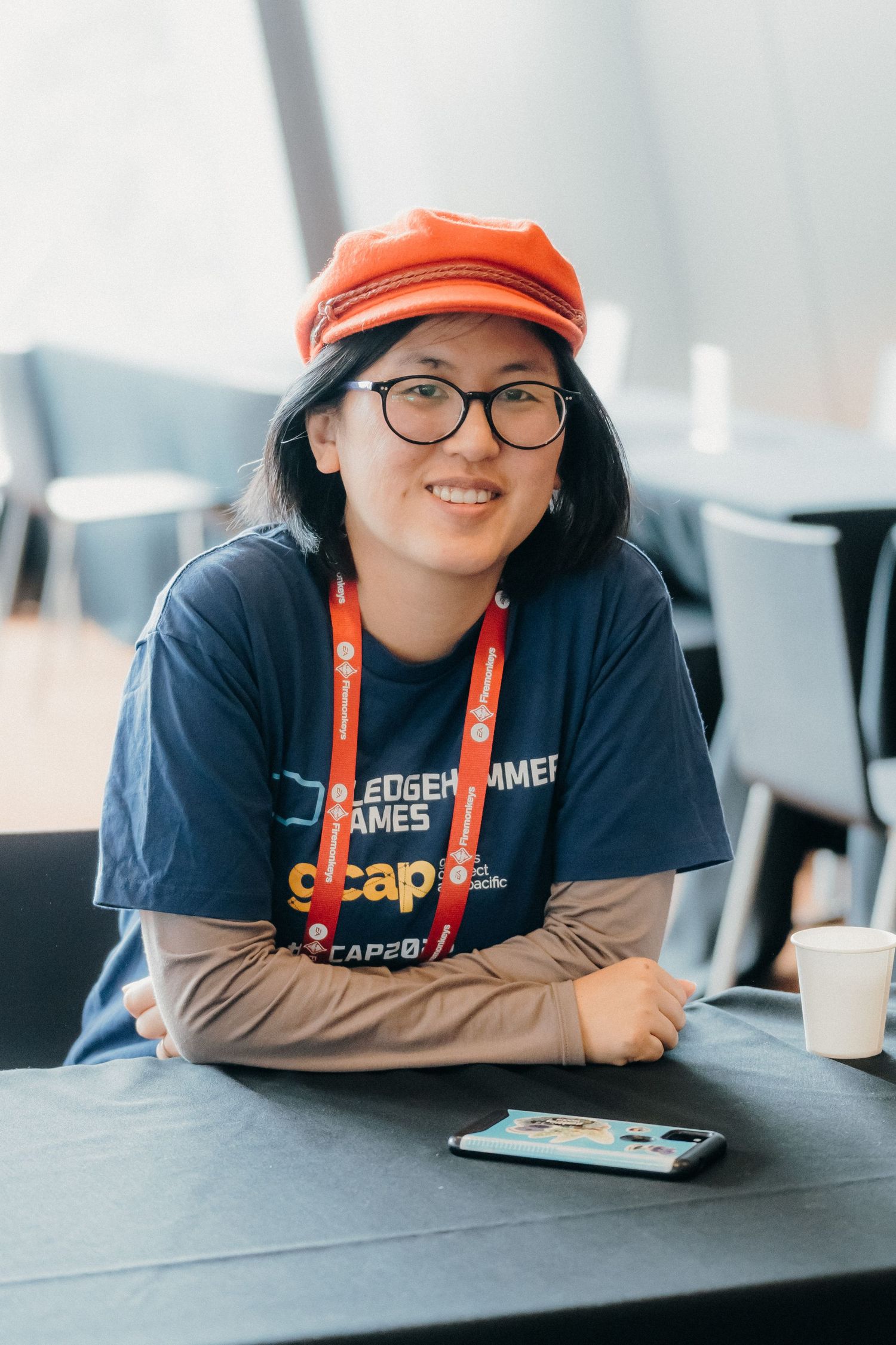 A person wearing glasses and an orange cap smiles while leaning on a gray table in an office setting.