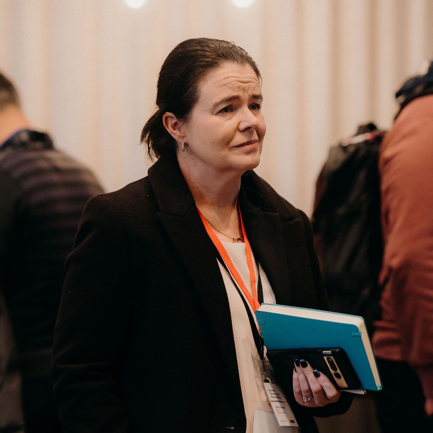Professional wearing a black coat and name badge holds folders while attending an indoor business event.