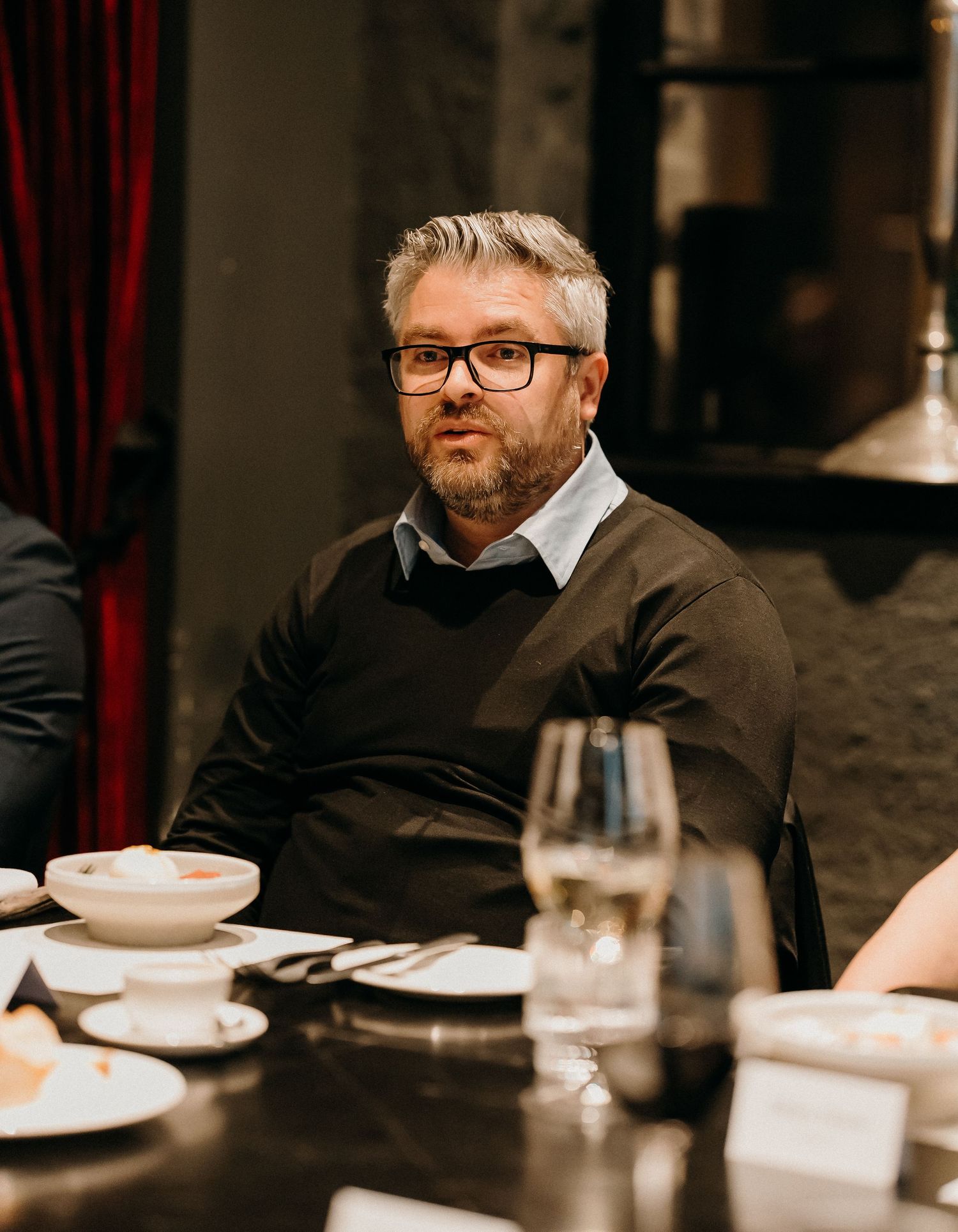 Person in glasses and brown sweater sitting at formal dining table with white tablecloth and place settings.