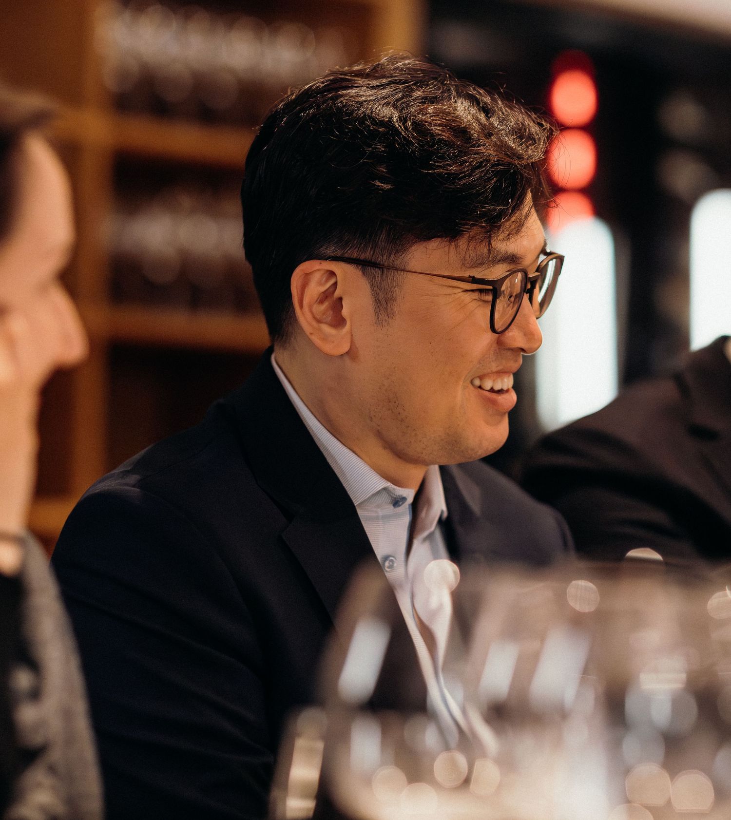 Professional wearing glasses and dark suit laughs during conversation in elegant restaurant setting.