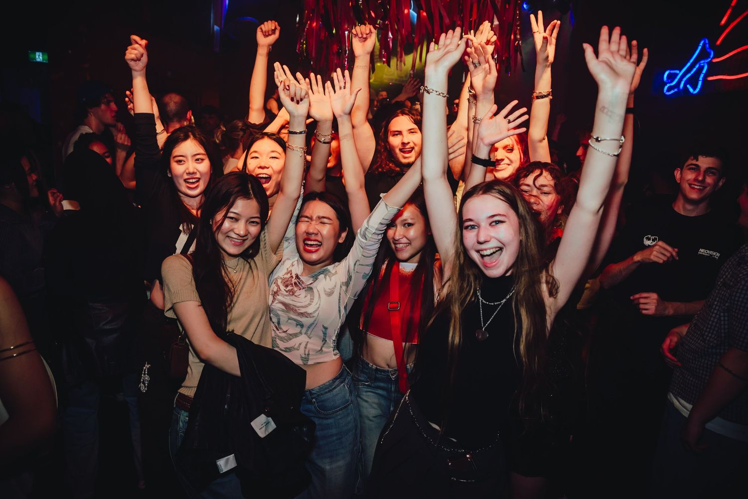 Crowd of energetic people dancing with raised hands at a nightclub with colorful lights and dark atmosphere.