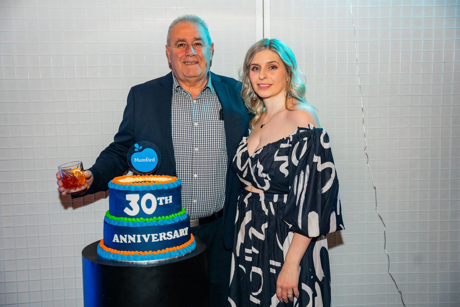 Two people in black and white outfits pose next to a large blue tiered anniversary cake at an event.