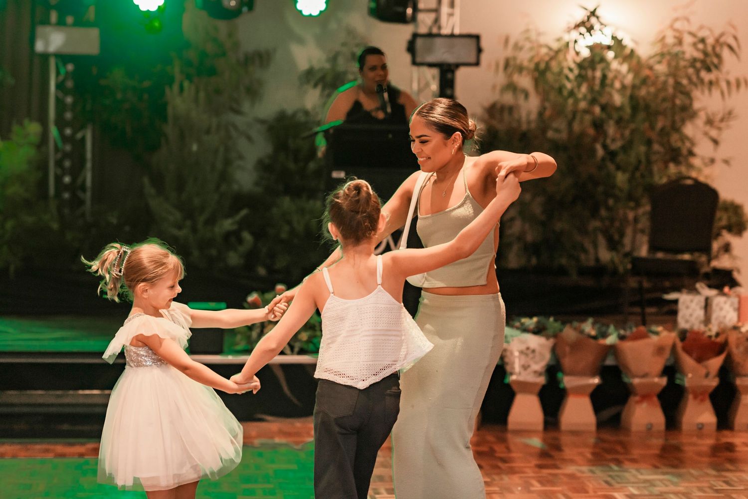 Dancers in white outfits spin together during an outdoor performance with green lighting and decorative plants in background.