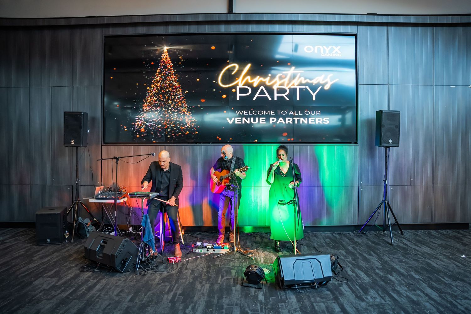 A band performs on stage at a Christmas party with colorful lighting and a decorated tree in the background.