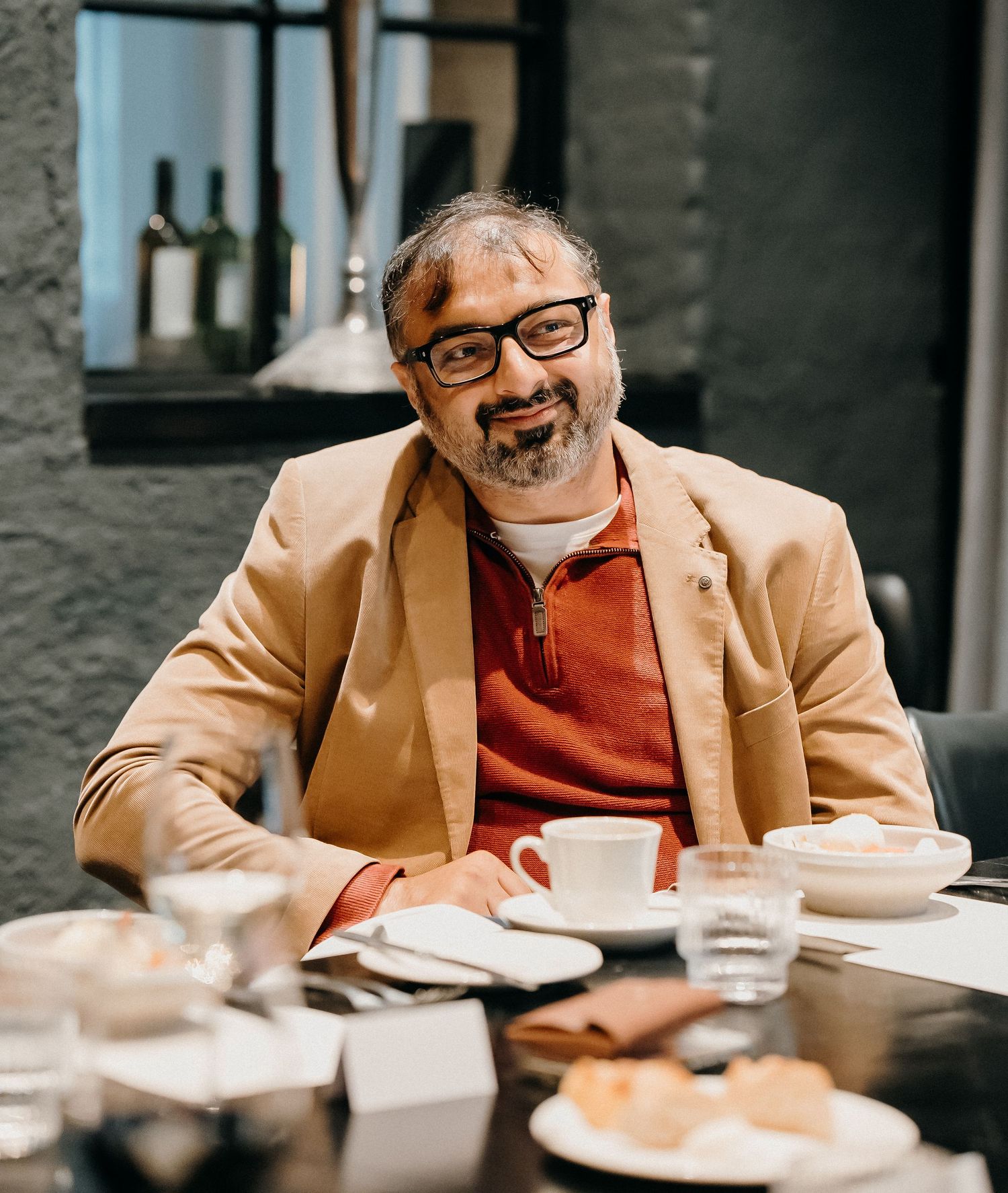 Person in glasses and tan blazer sits at restaurant table with food and drinks, smiling while looking down at plates.