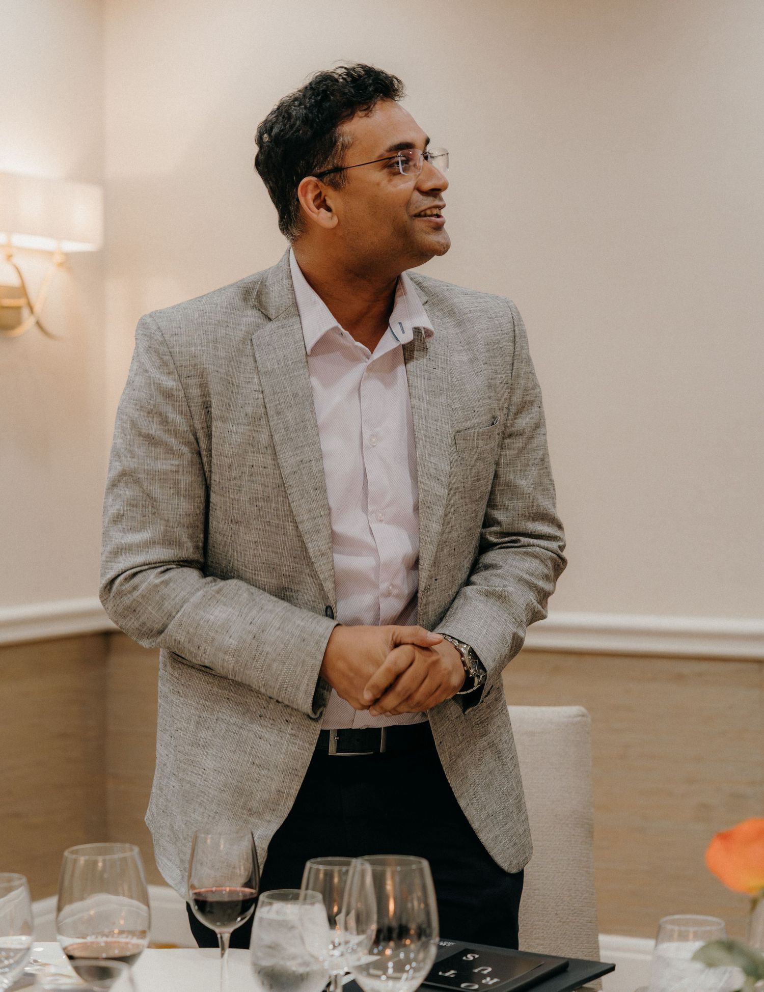 A person in a gray blazer and white shirt sits at a dining table with wine glasses and place settings.