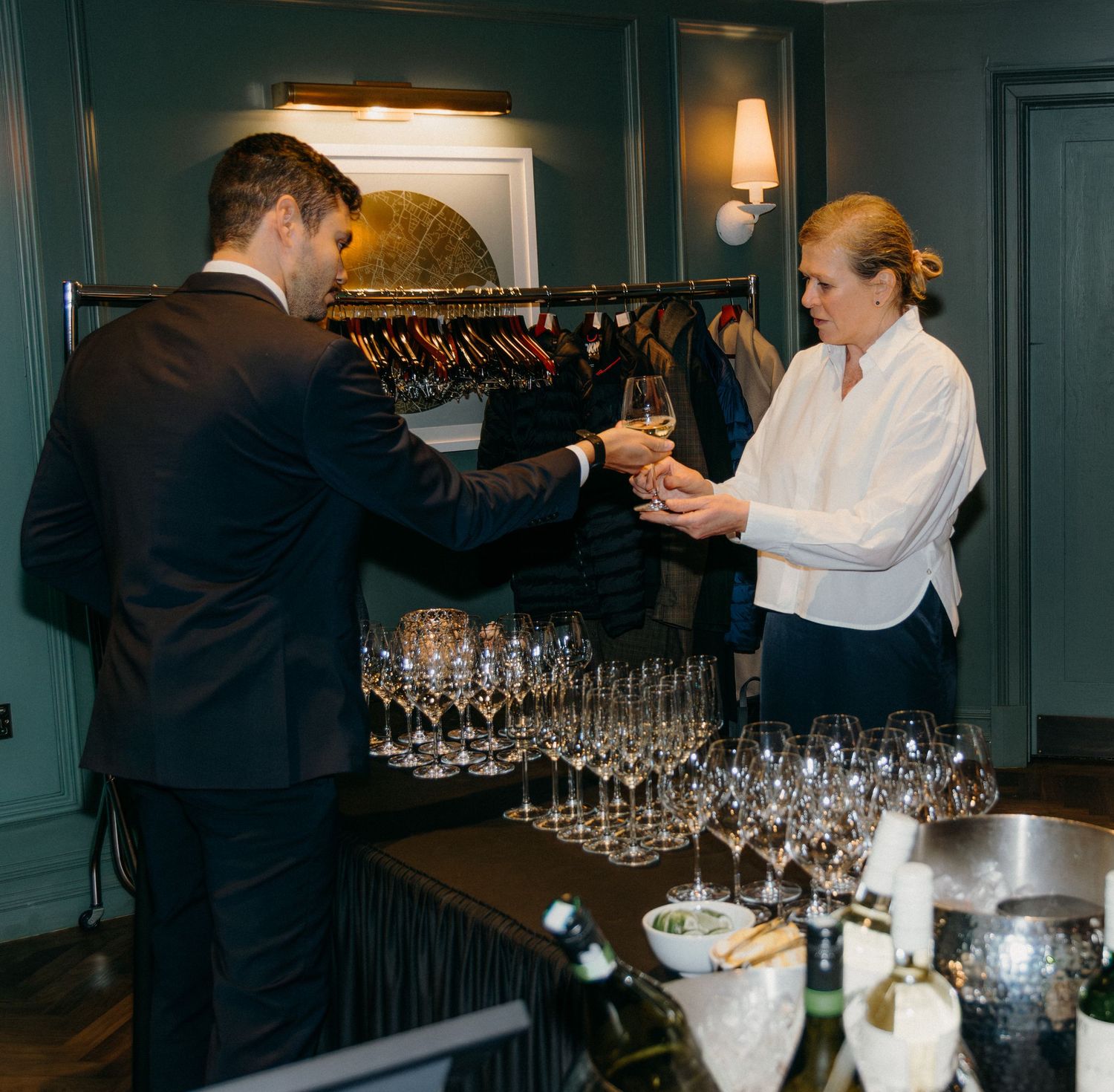 Bartenders setting up rows of wine glasses at an elegant event bar with dark teal walls and ambient lighting.