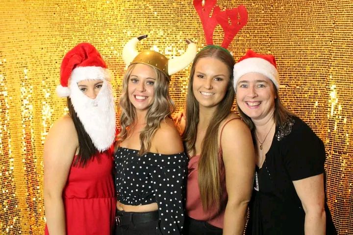 Group poses together in holiday attire with Santa hats and reindeer antlers against a gold sequin backdrop.