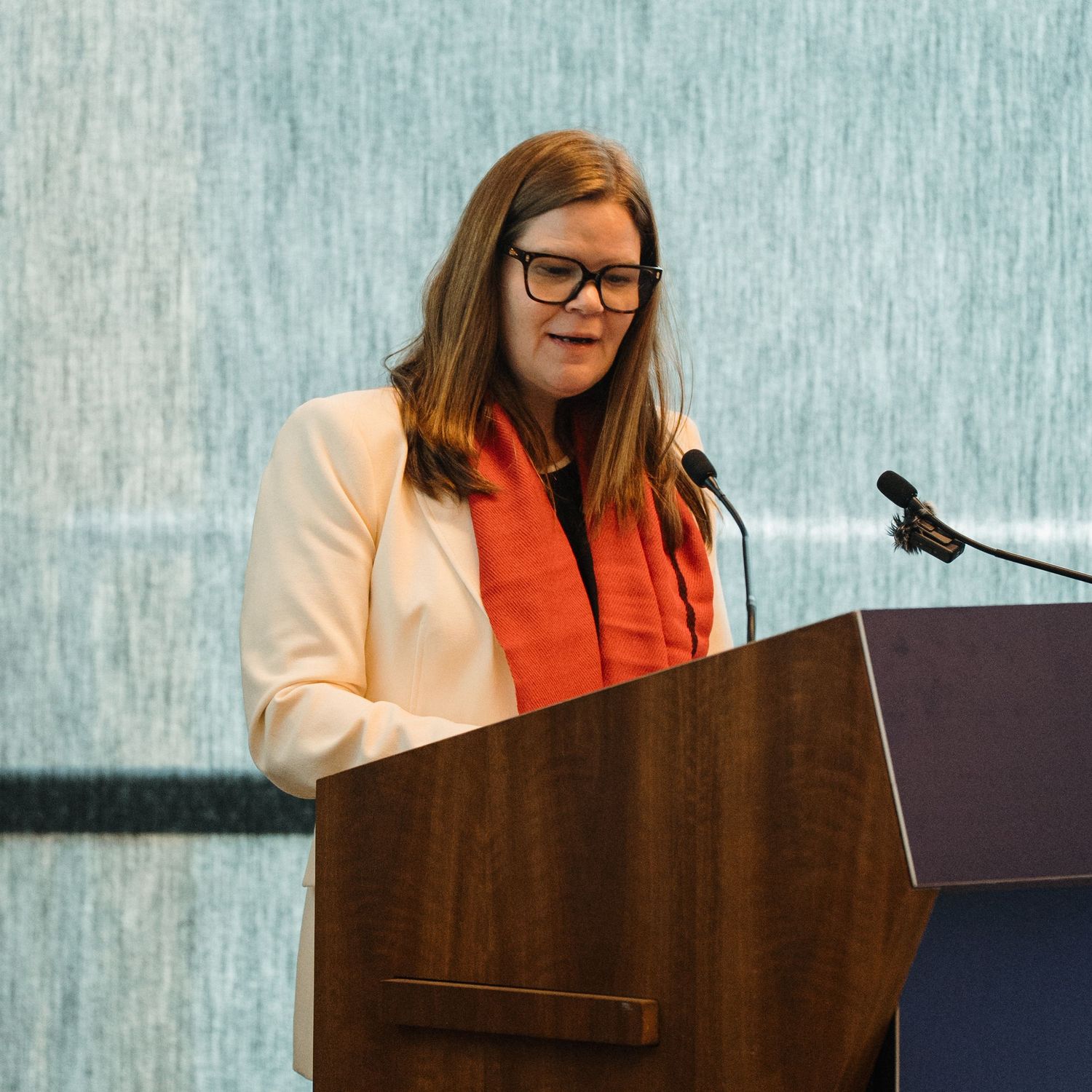 Speaker at wooden podium wearing glasses and white blazer with coral top presenting at professional conference event.