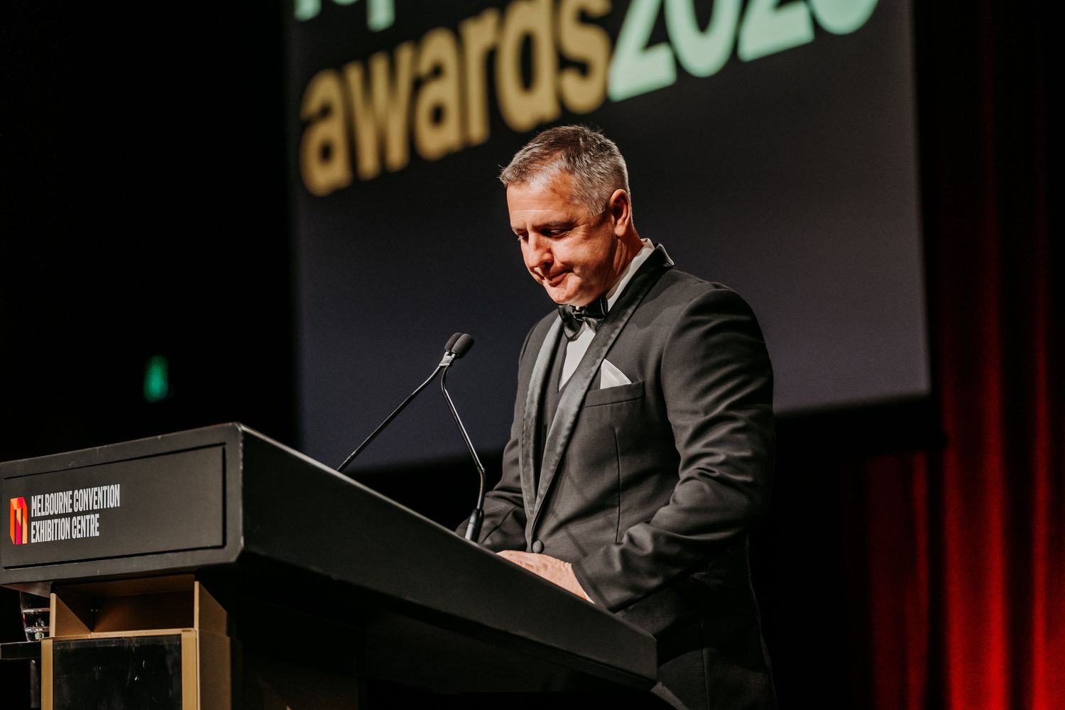 A speaker stands at a podium giving a presentation at an awards ceremony with red curtains in the background.