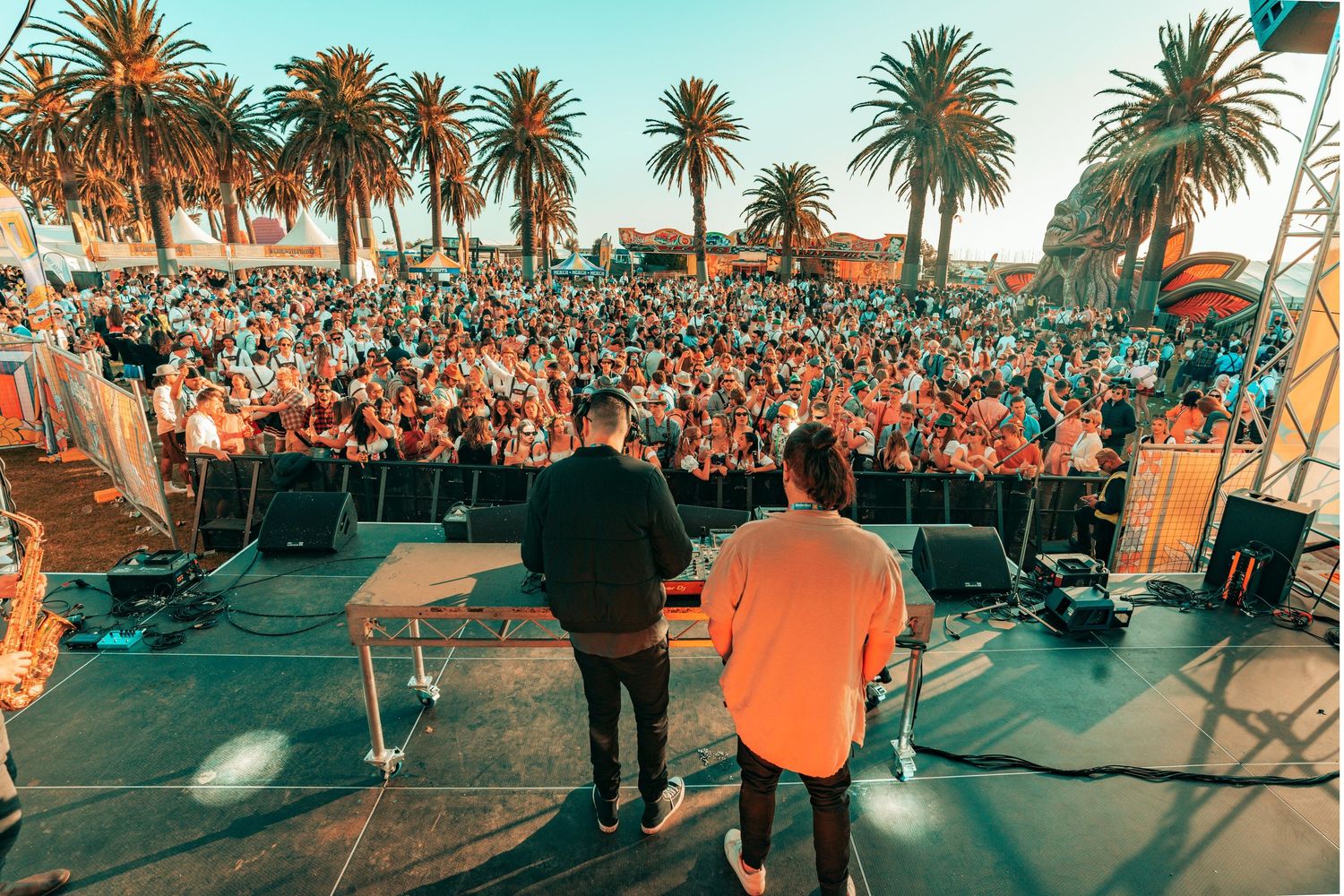 Large outdoor crowd gathered at a beachside concert venue with palm trees during sunset.