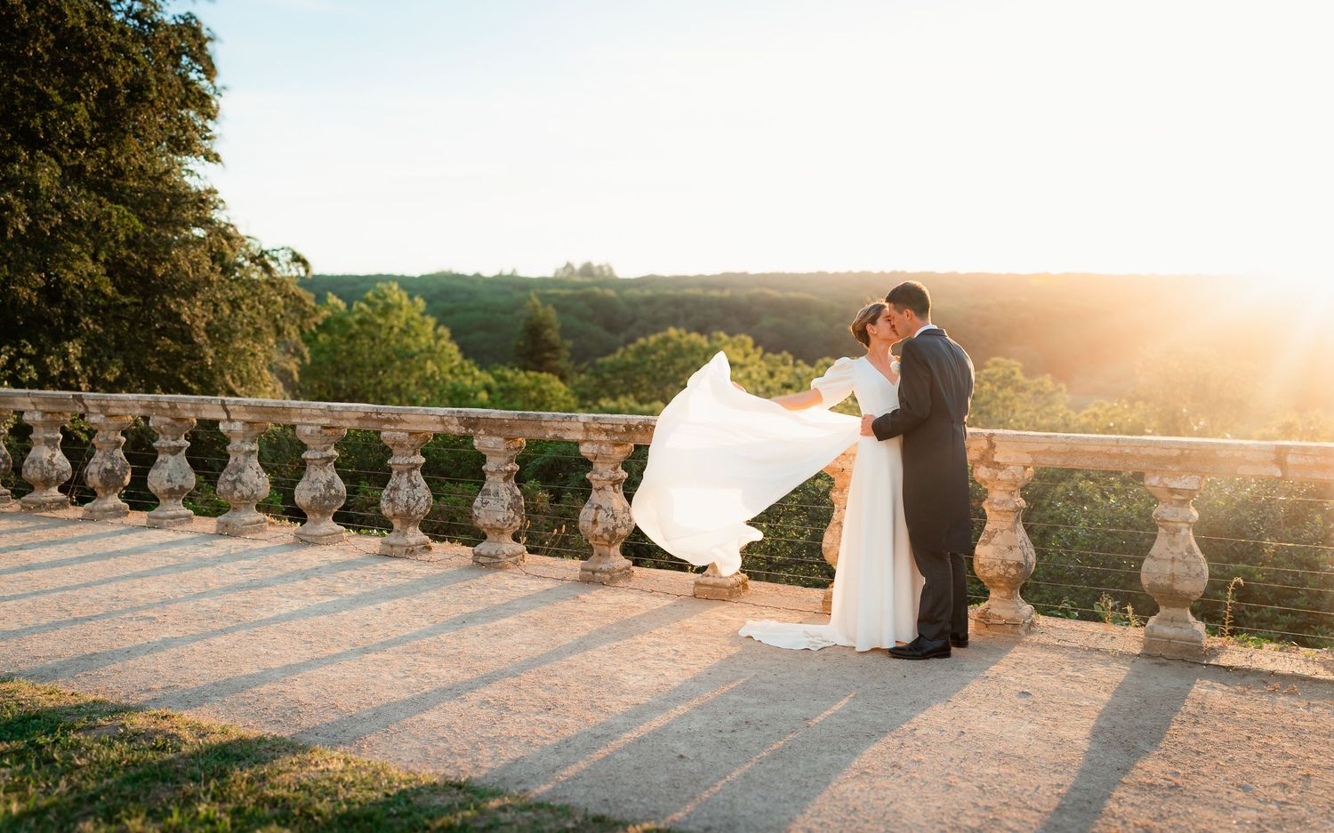 Bride and groom share romantic moment on sunlit stone terrace with scenic countryside views at golden hour.
