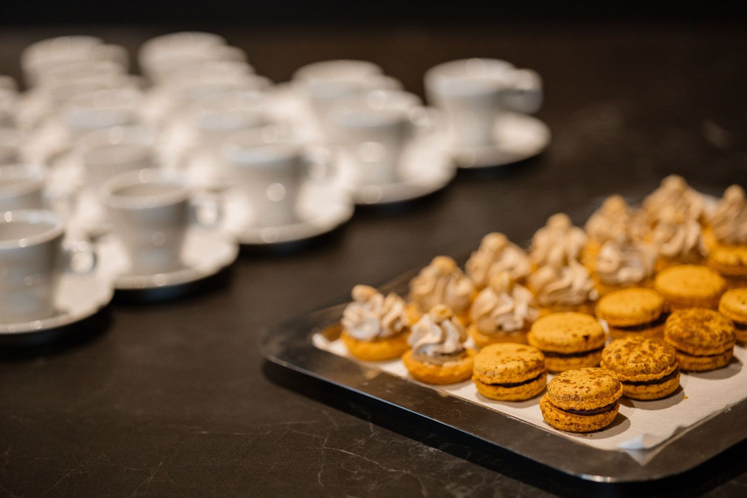 Rows of golden macarons and white coffee cups arranged on dark serving tray.