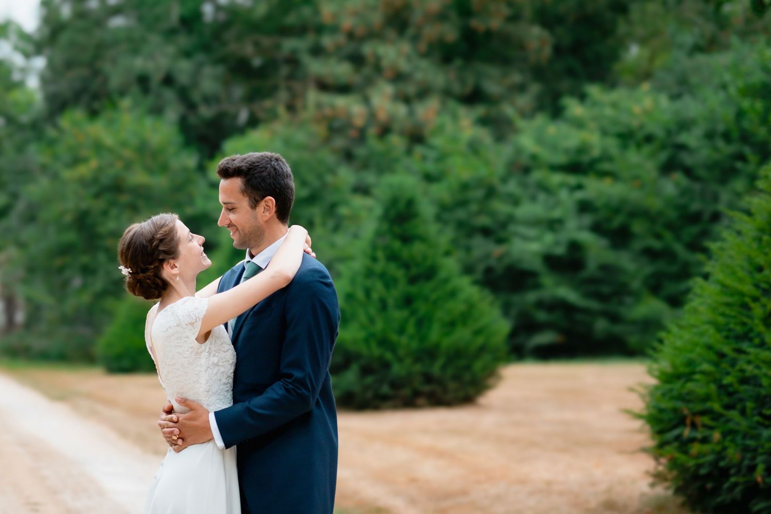 Happy newlywed couple embraces outdoors with lush green trees and garden path in background.