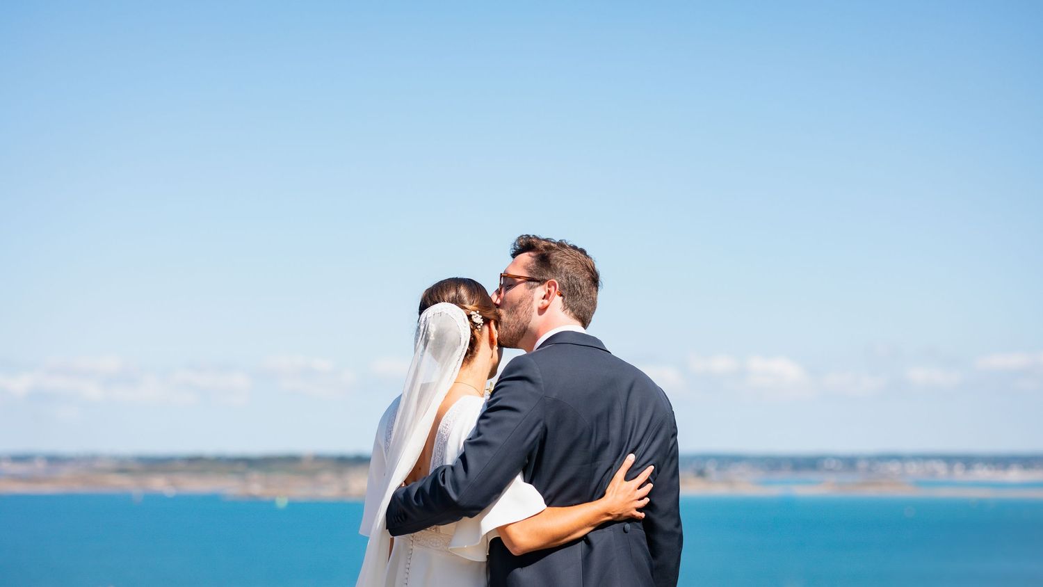 A couple embraces on a cliff overlooking the ocean on a sunny day with blue skies and water in the background.