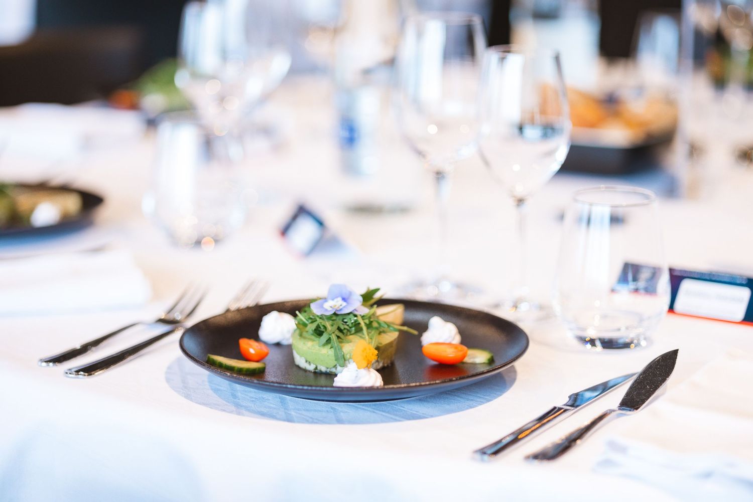 Gourmet plated dish with colorful garnishes served on dark plate with white tablecloth.