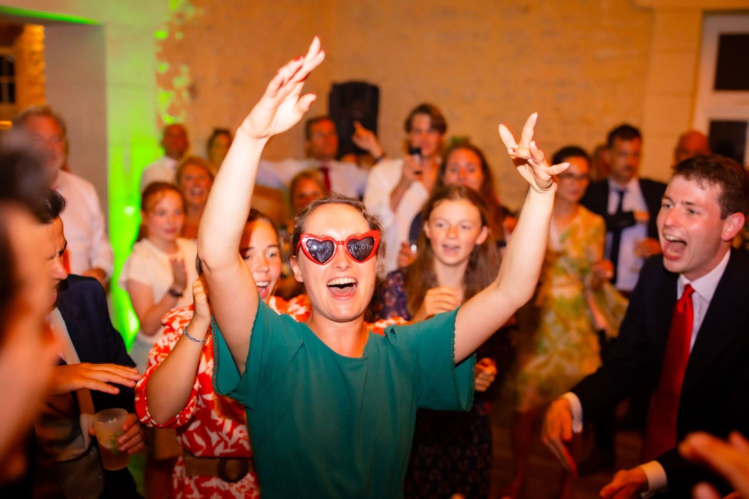 Wedding guests dancing enthusiastically at a reception with colorful lighting and brick wall backdrop.