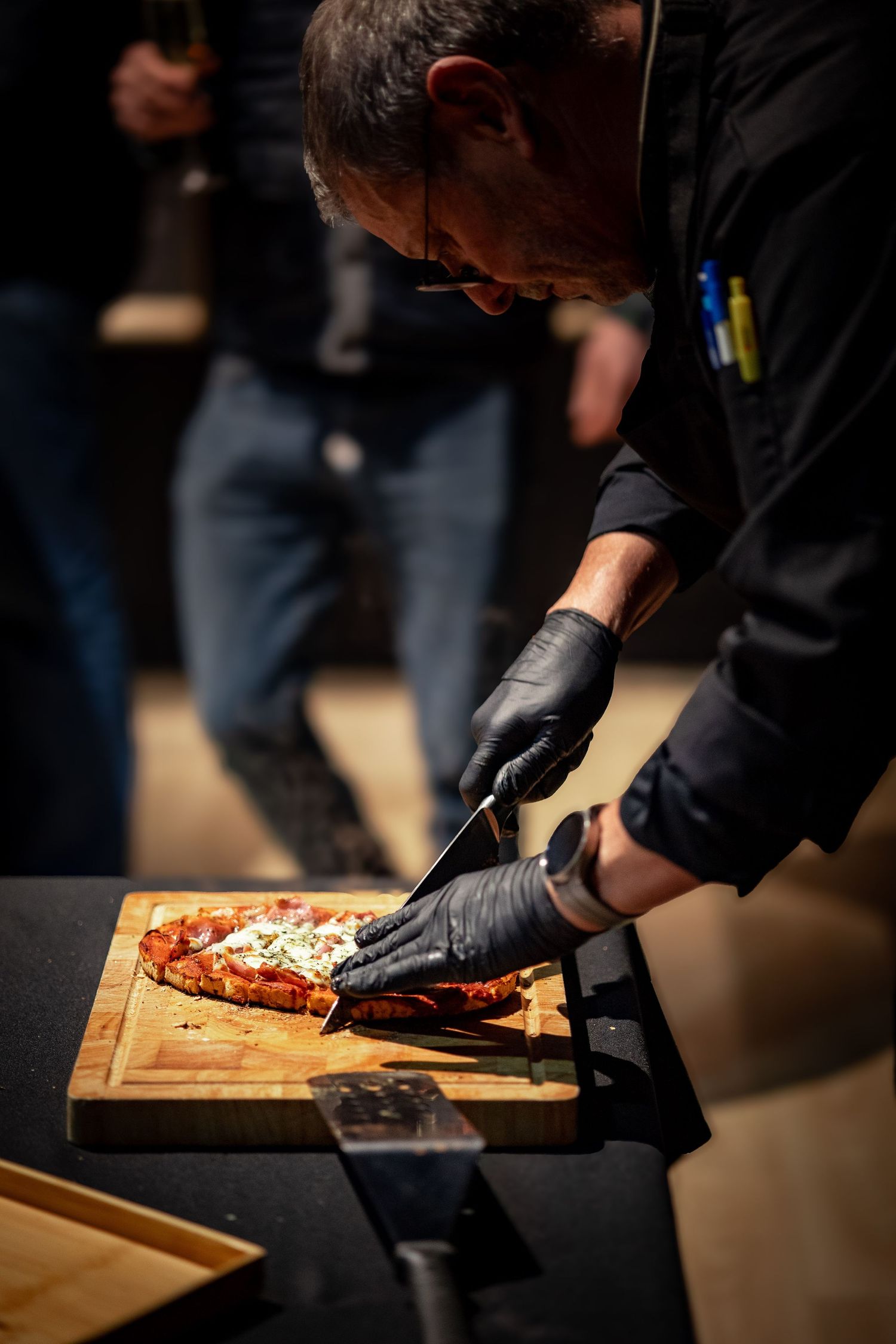 Chef with black gloves cutting pizza on wooden board.