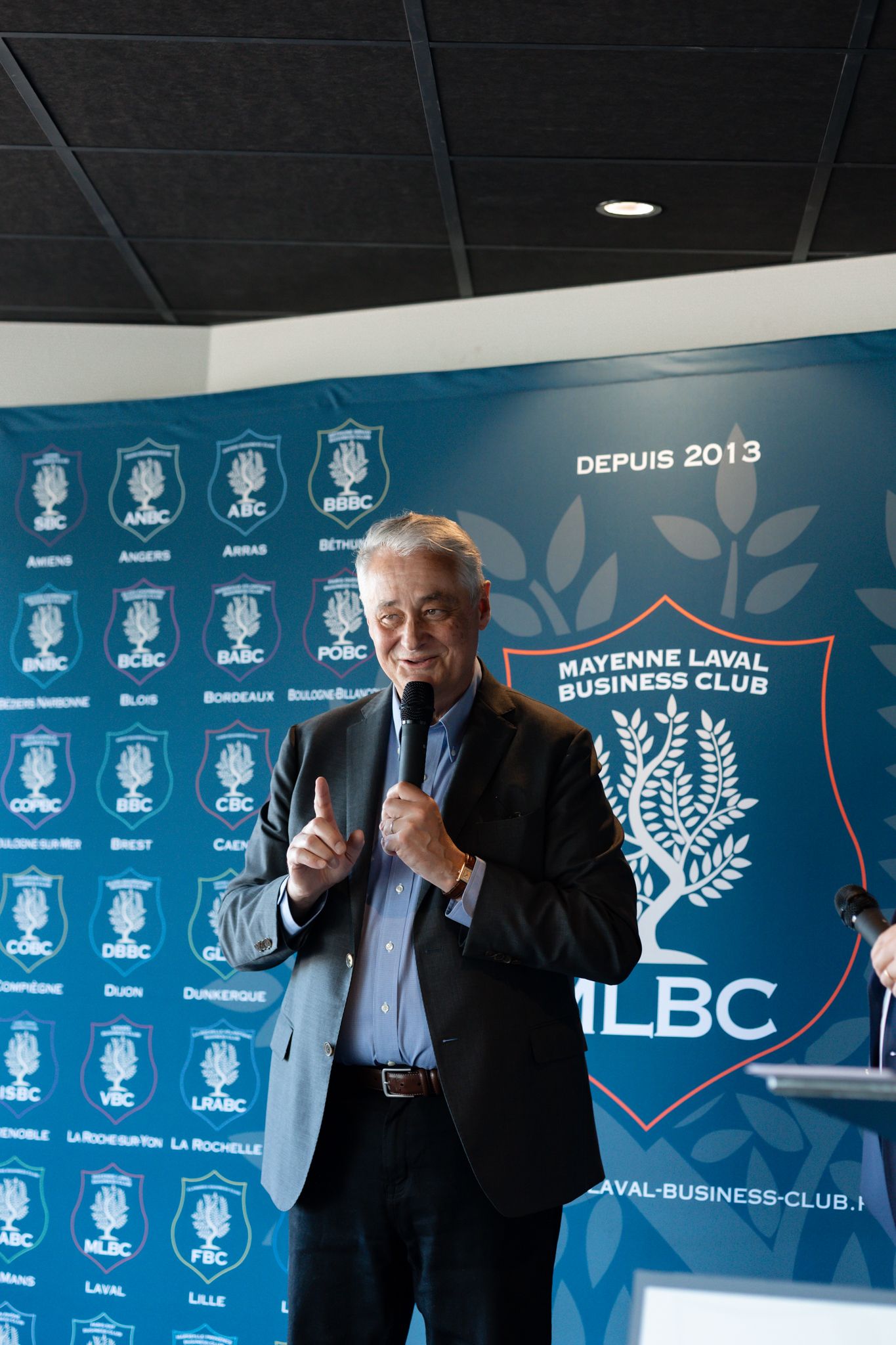 A speaker presents at a podium in front of a blue backdrop with the Mayenne Laval Business Club (MLBC) logo and branding.