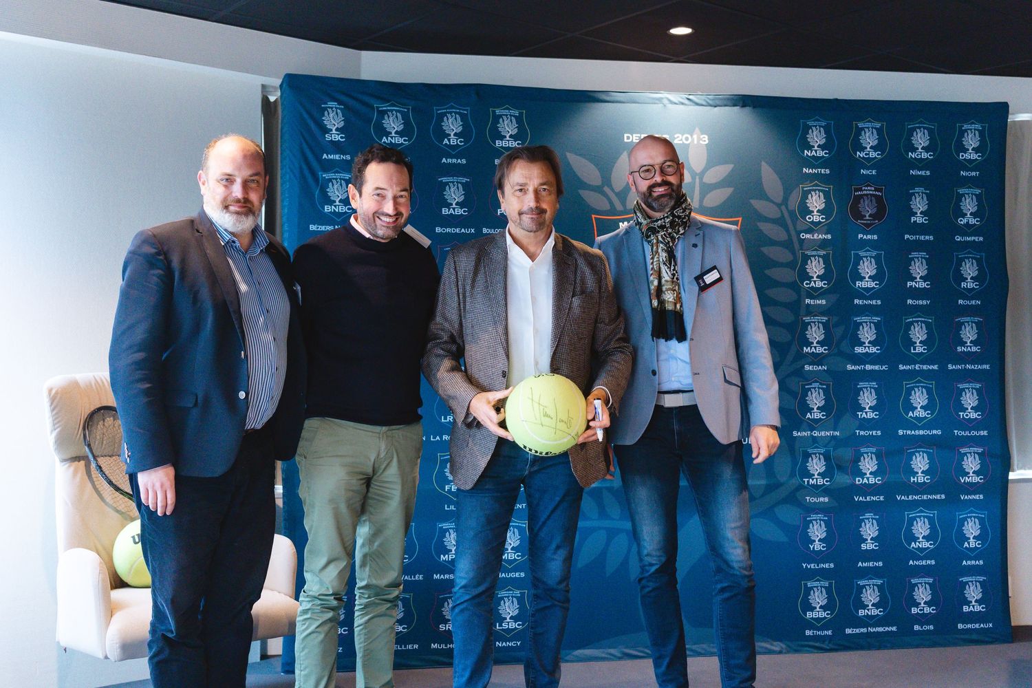 Four men standing in front of a blue backdrop with sports logos holding a tennis ball.