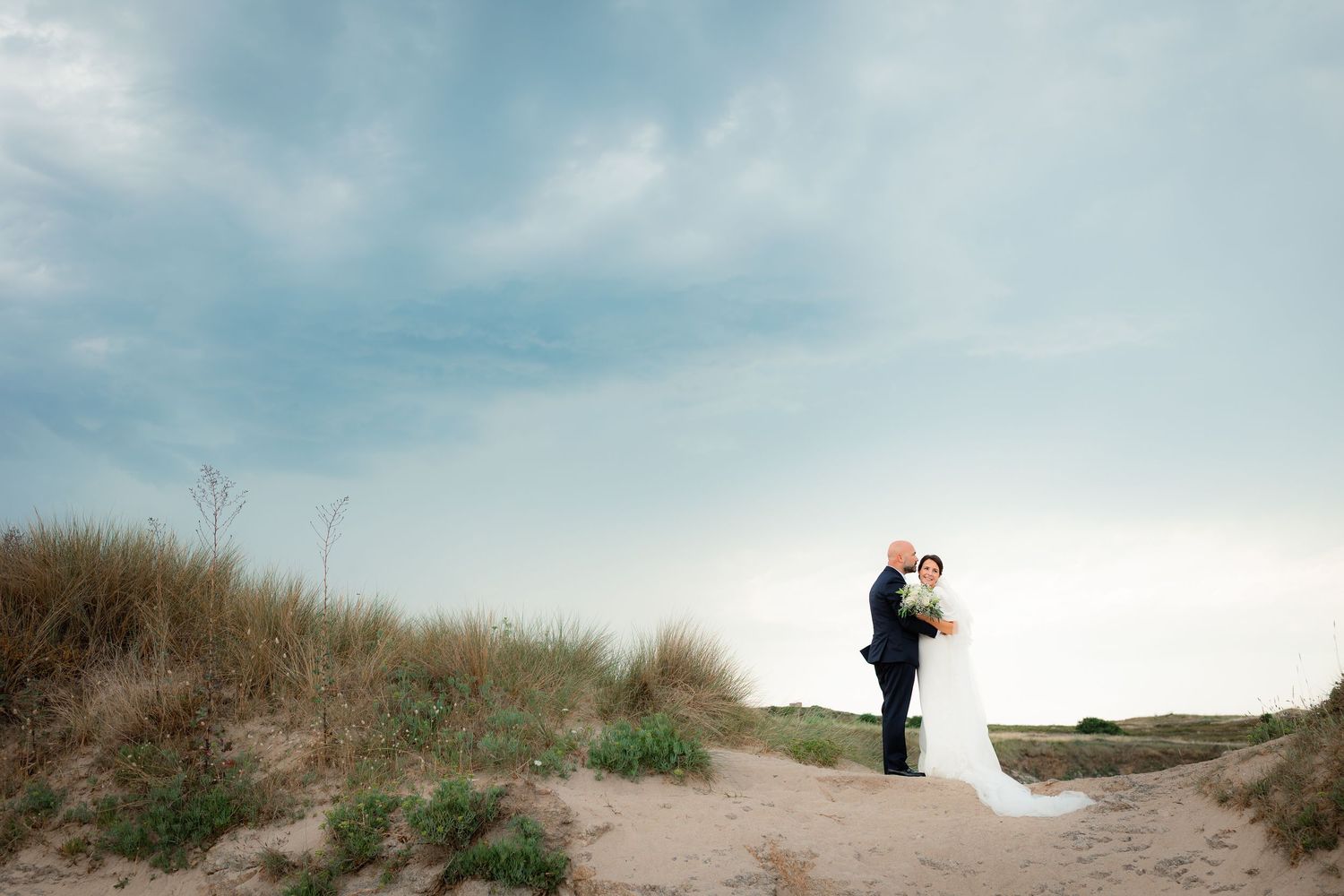 Bride and groom embracing on sandy beach dunes under a dramatic cloudy sky at their outdoor wedding.