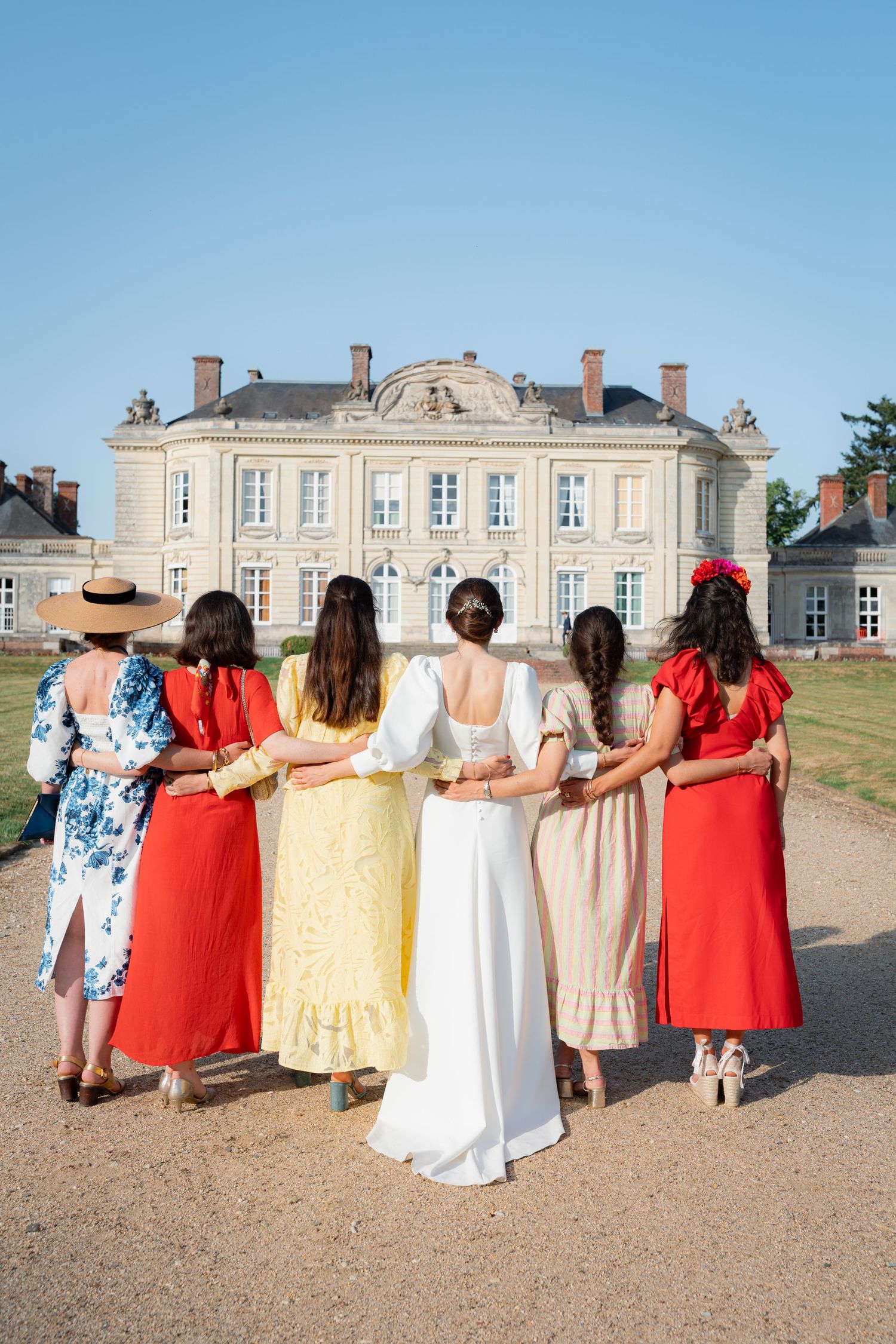 Bride and bridesmaids in colorful dresses stand arm in arm facing a grand French chateau on a sunny day.