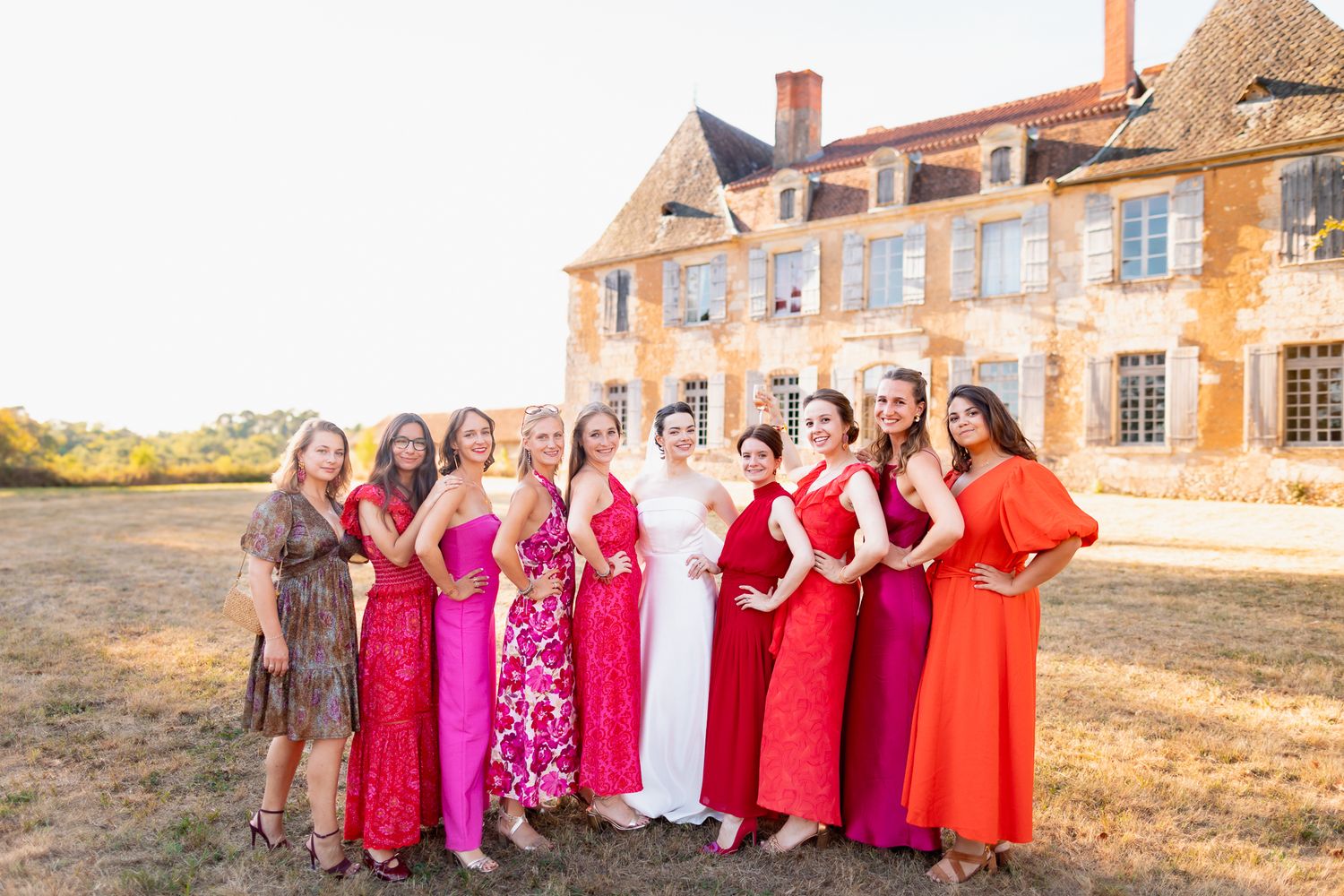 Bride in white gown poses with bridesmaids in pink and red dresses outside a French chateau on a sunny day.
