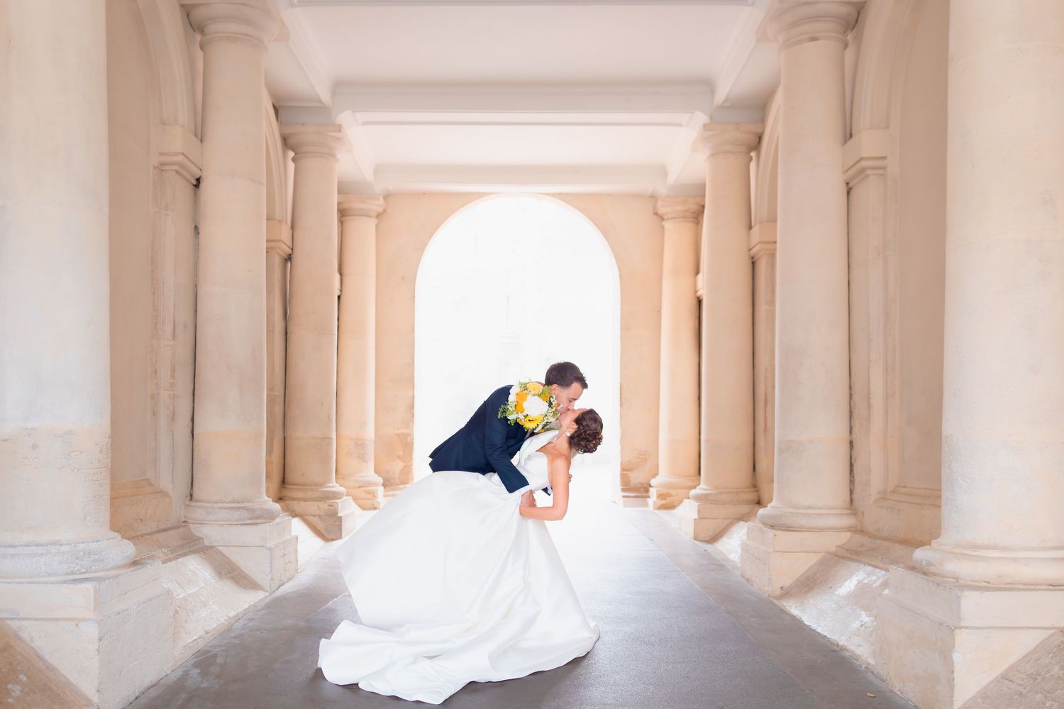 An elegant wedding couple shares a romantic moment in a grand colonnade with white architectural pillars and arches.