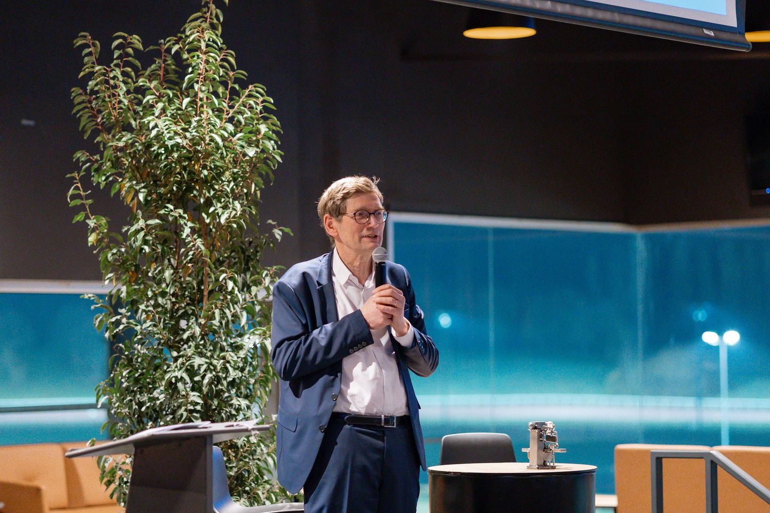 Speaker presenting at a podium with a decorative plant in a modern venue setting.