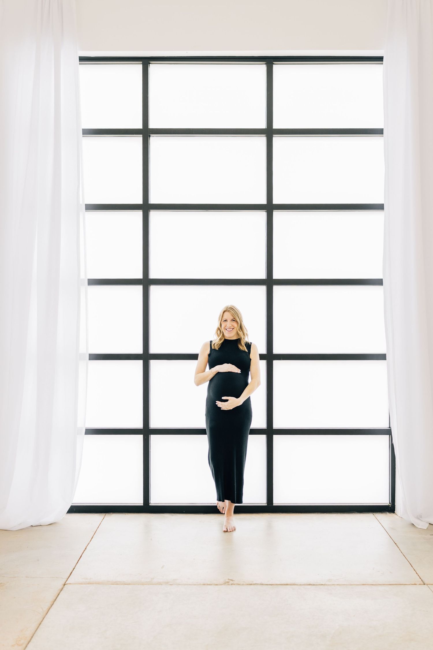 Figure in a black outfit stands in front of a modern geometric window with white curtains in a bright minimal space.