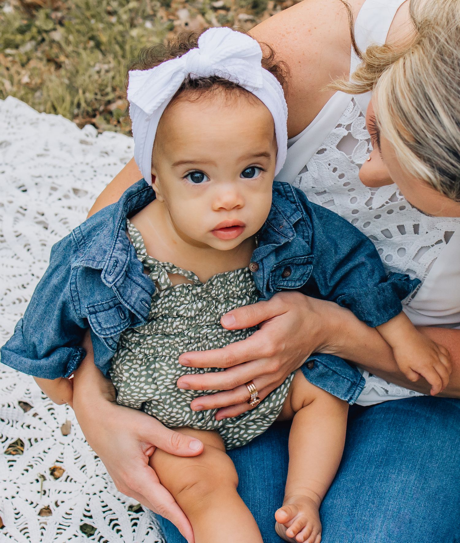 Baby wearing blue denim top and floral outfit sits on white blanket in grass while being held.