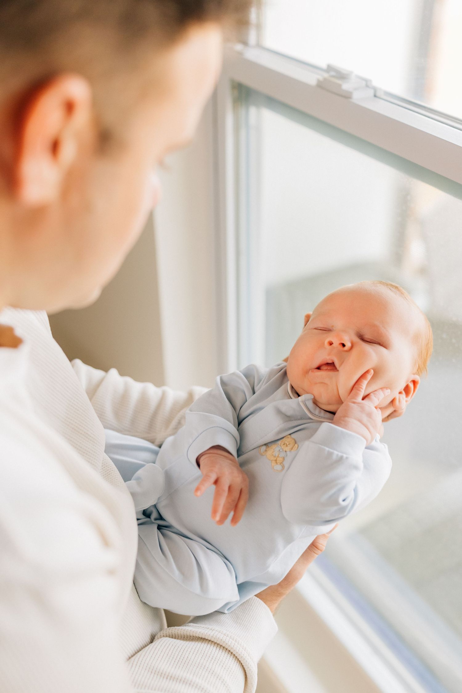 A peaceful sleeping newborn baby wearing light blue sleepwear is cradled near a bright window.