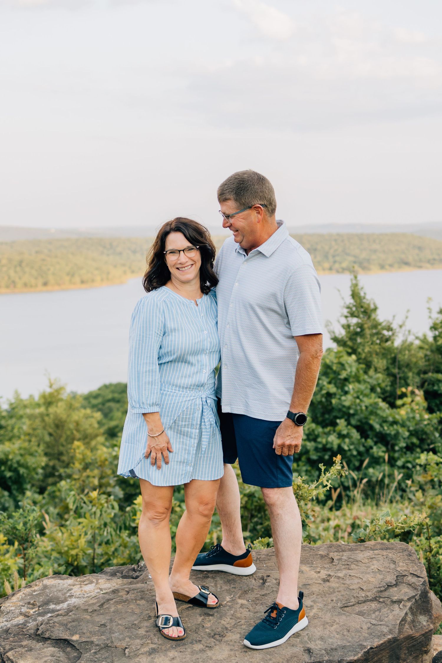 A couple stands together on a rocky overlook with a scenic lake view in the background.