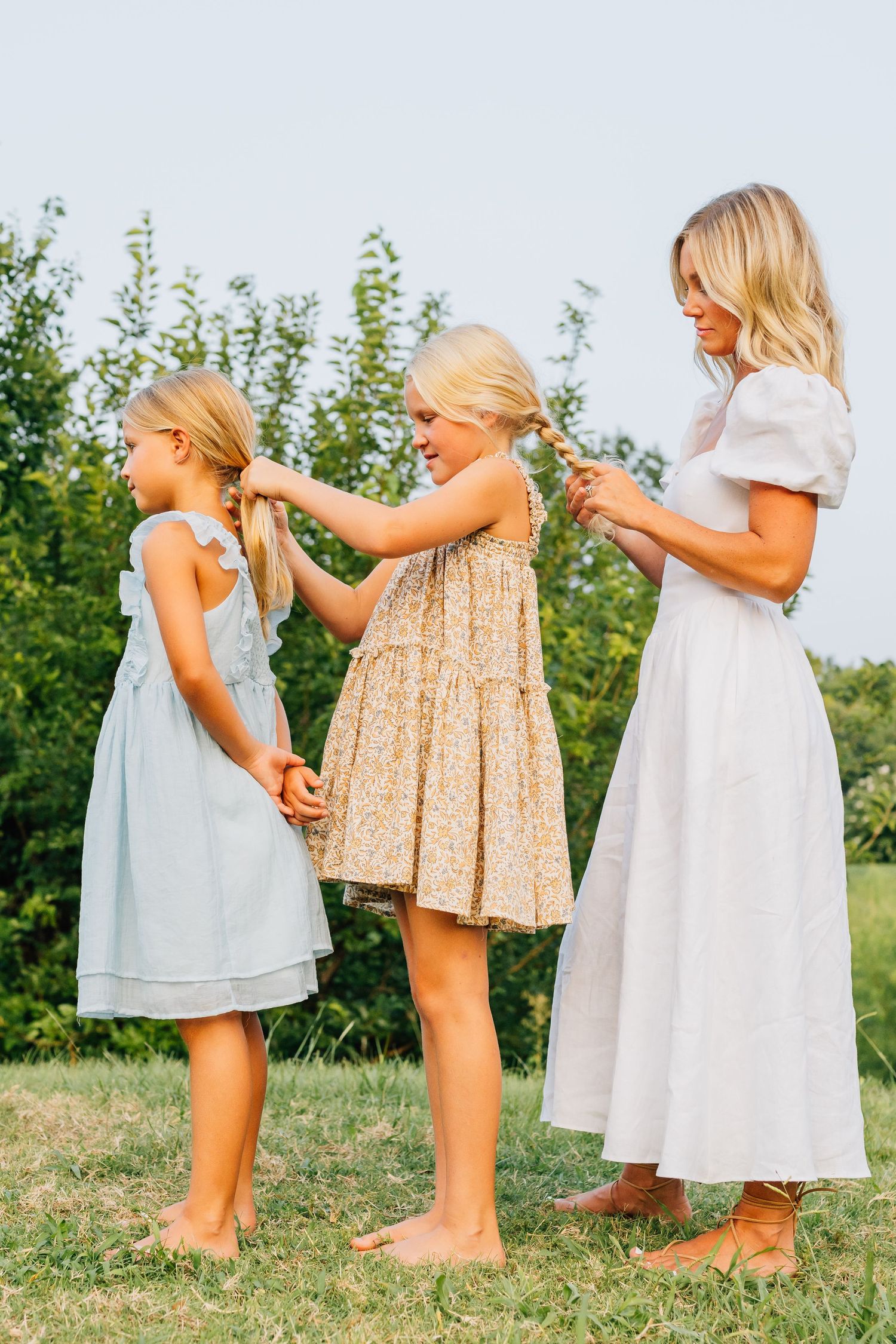 Three people wearing summer dresses stand in line braiding each other's hair in a sunny garden.