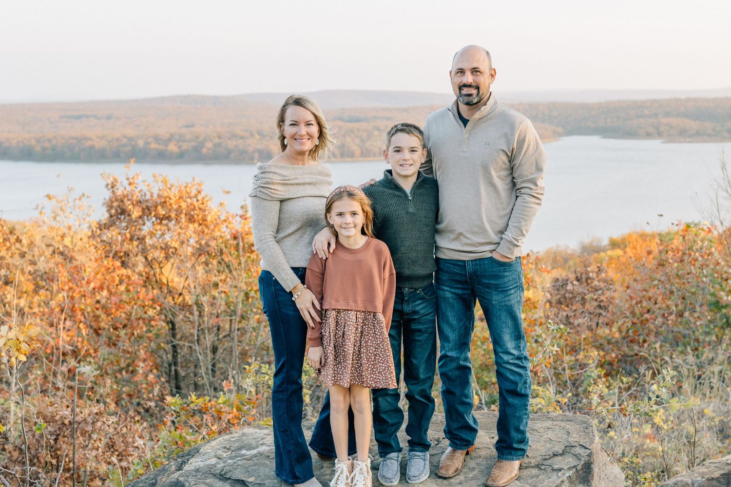 A family of four poses together on a hilltop with autumn foliage and a lake in the background.