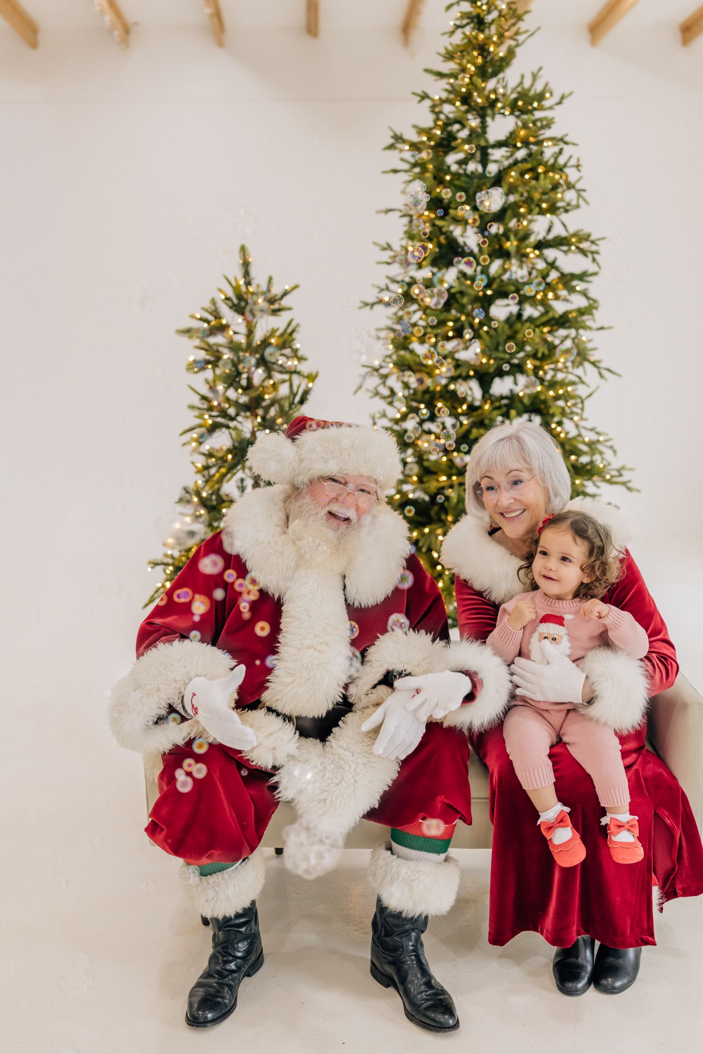 A toddler sits with Santa Claus and Mrs. Claus in red velvet outfits in front of lit Christmas trees.