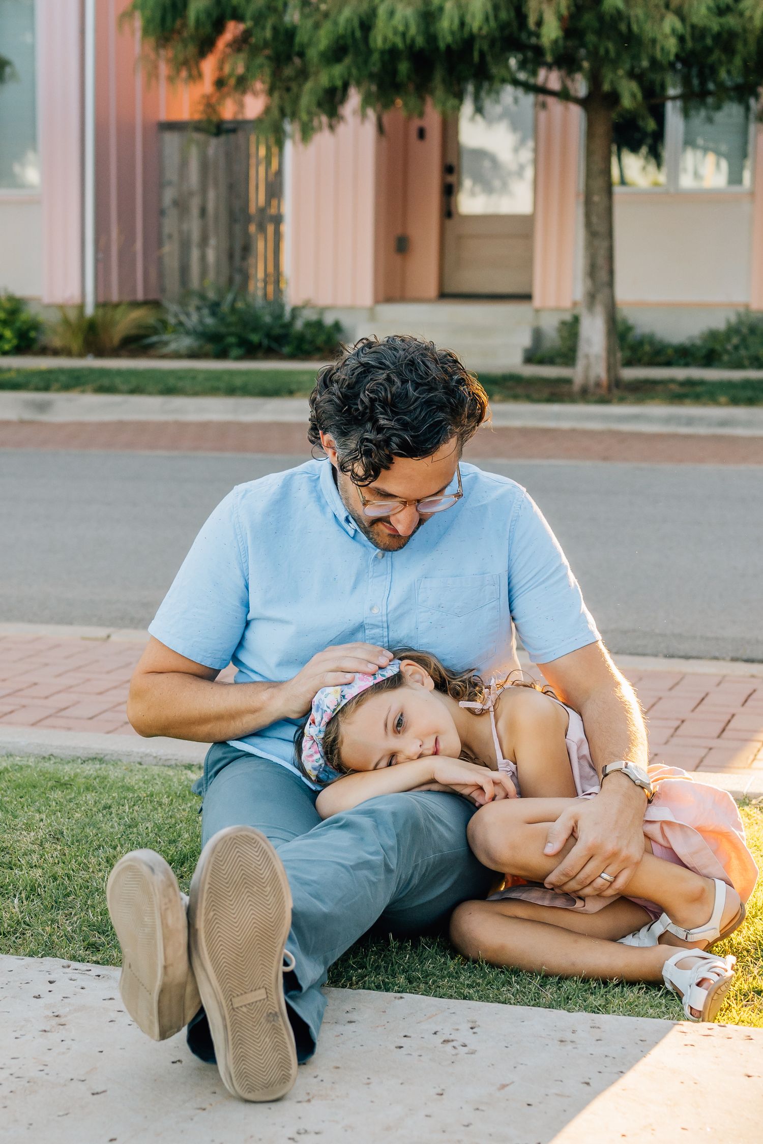 A father and child share a tender moment while resting together on a sidewalk near grass and trees.