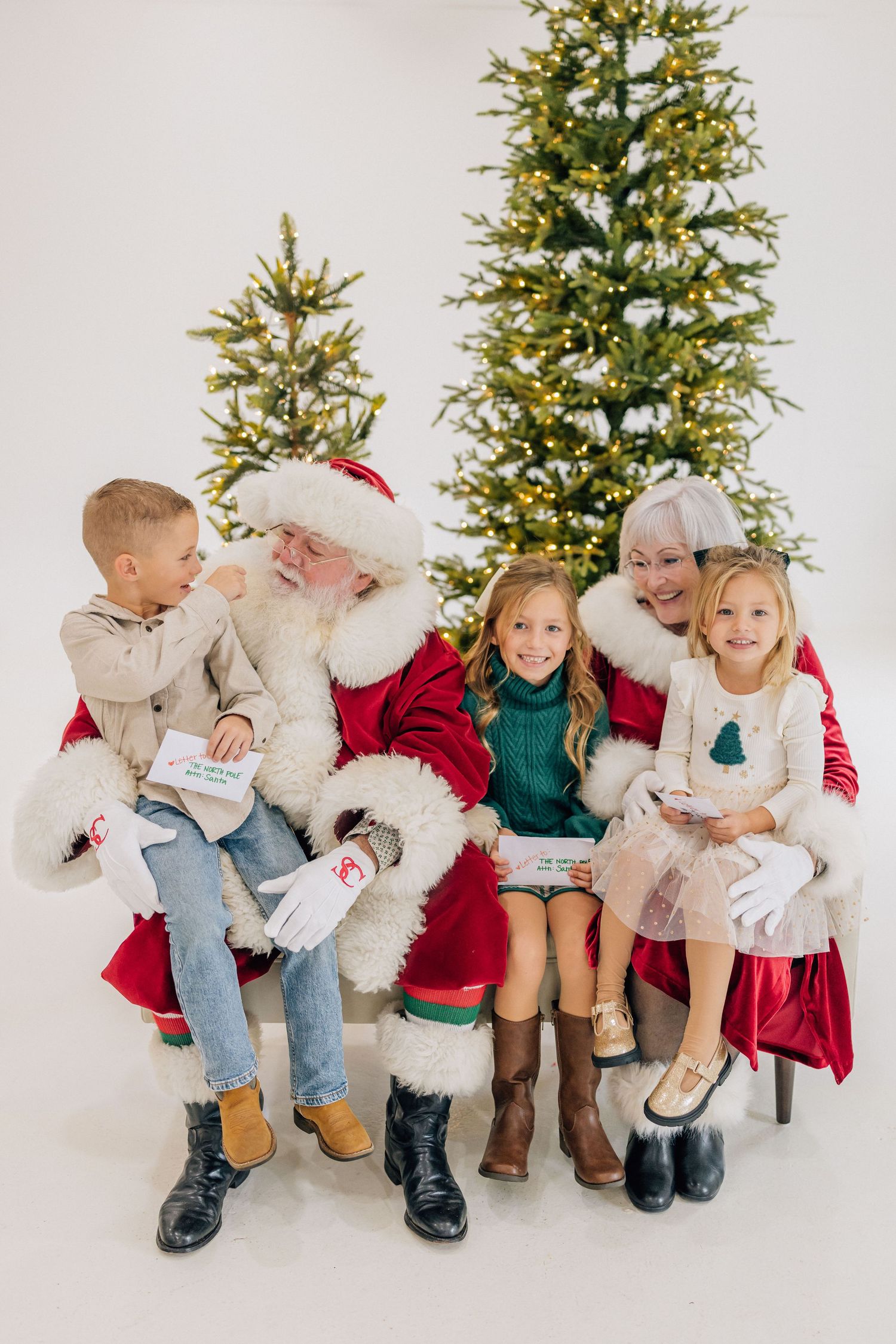 Santa and Mrs. Claus sit with three children in front of lit Christmas trees in a bright holiday photo.