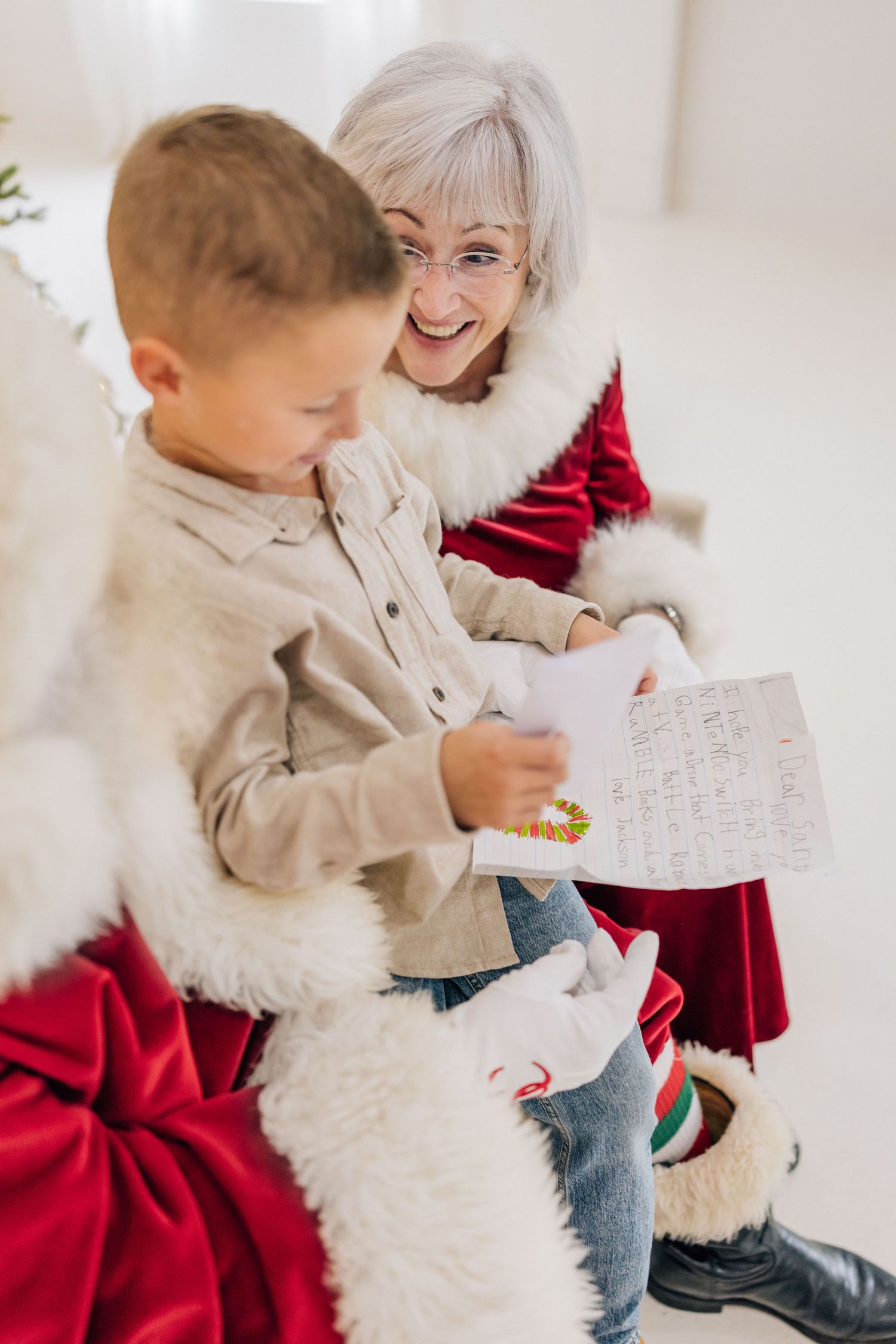 Mrs. Claus in red suit reads Christmas wish list with excited young boy sitting on her lap.