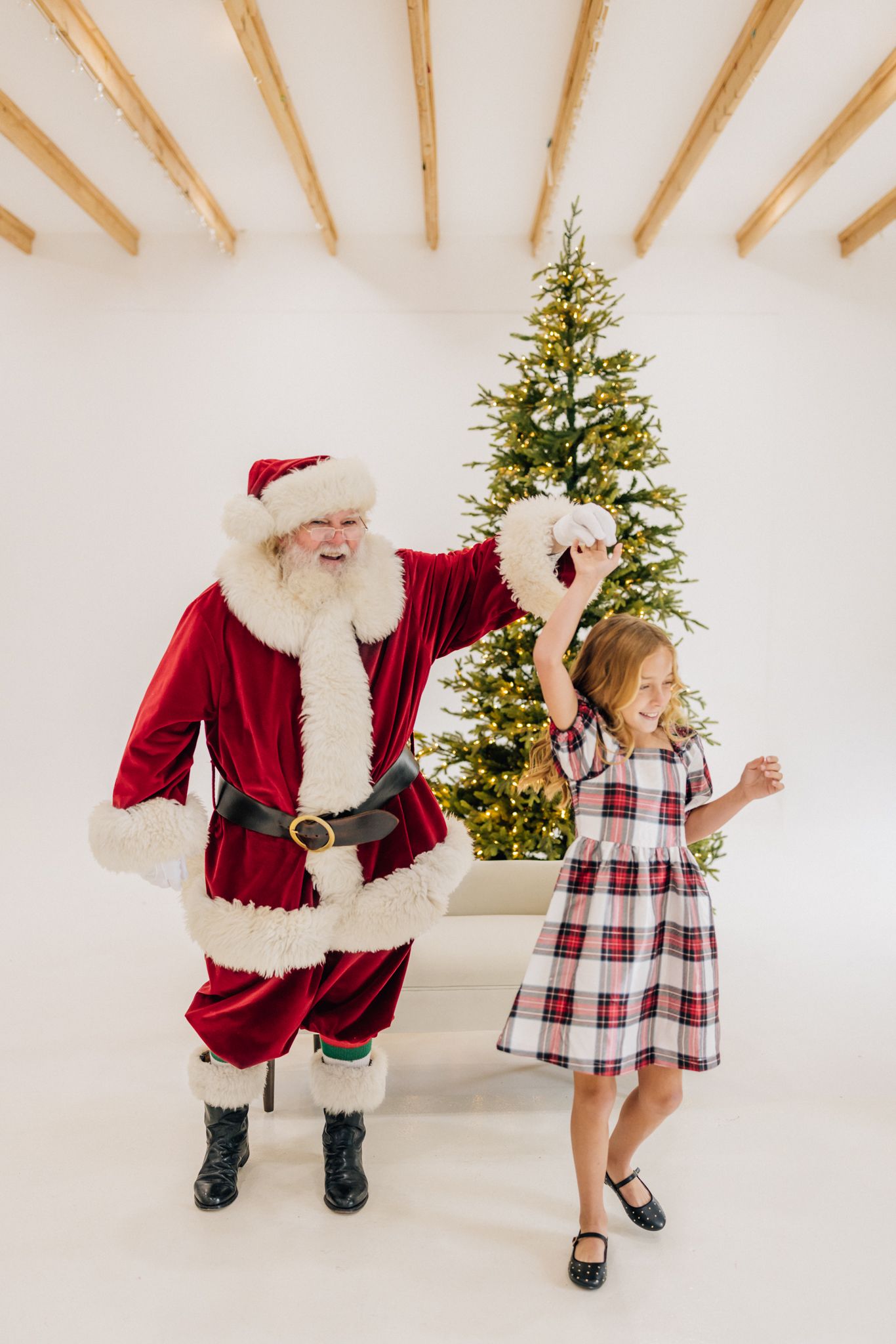 Santa Claus and a young girl in plaid dress dancing joyfully in front of a decorated Christmas tree.
