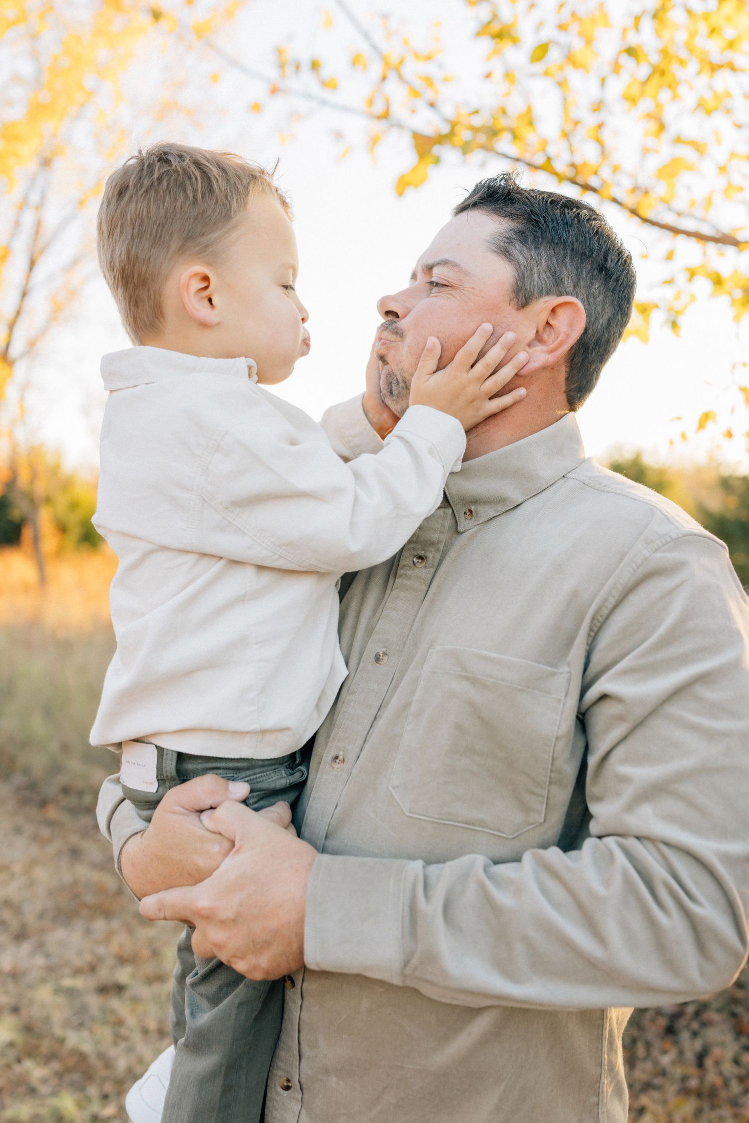 A tender moment captured outdoors in autumn as a toddler in white reaches to touch an adult's face.