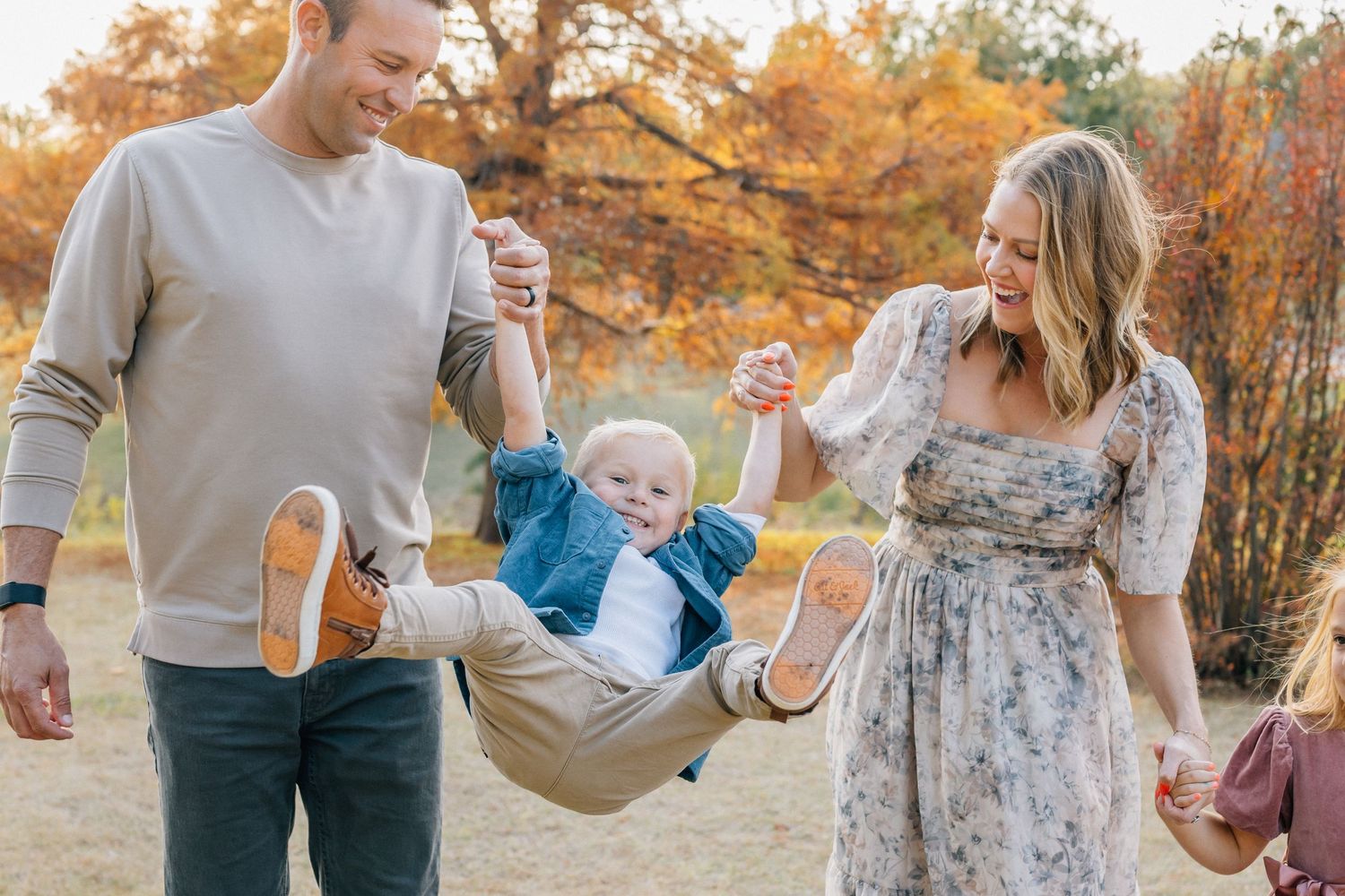 Family enjoying autumn day together swinging child between them on wooded path with colorful fall foliage.