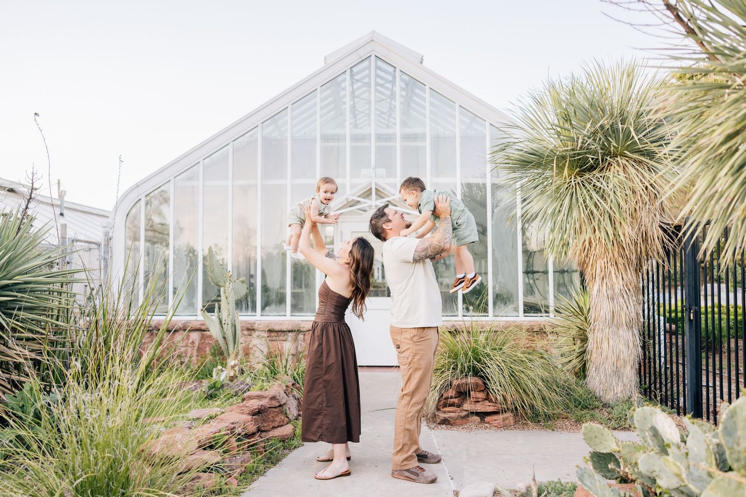 A family enjoys a tender moment together outside a glass greenhouse surrounded by desert plants and palm trees.