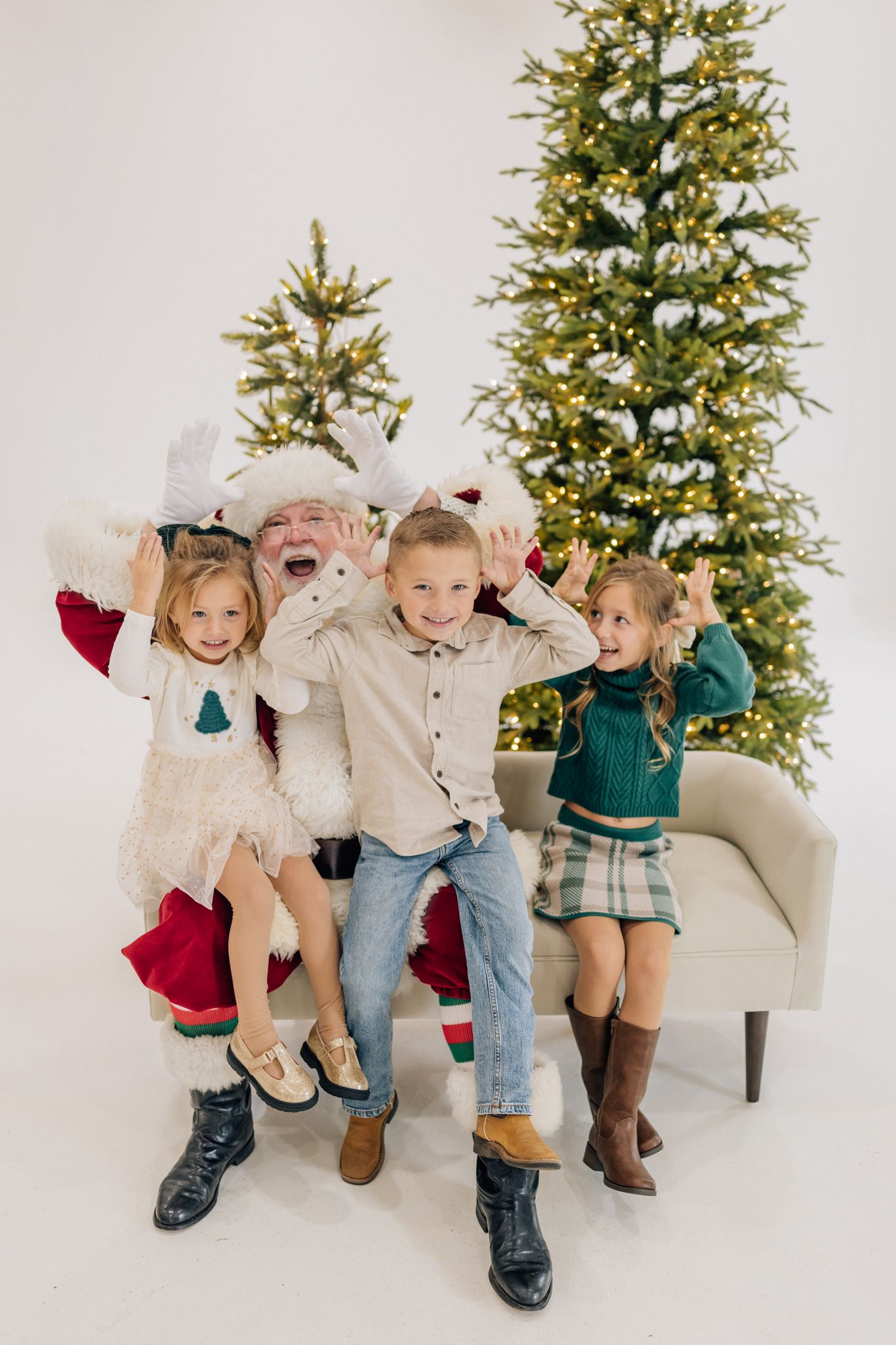 Three children pose playfully with Santa Claus in front of lit Christmas trees in a bright white studio setting.