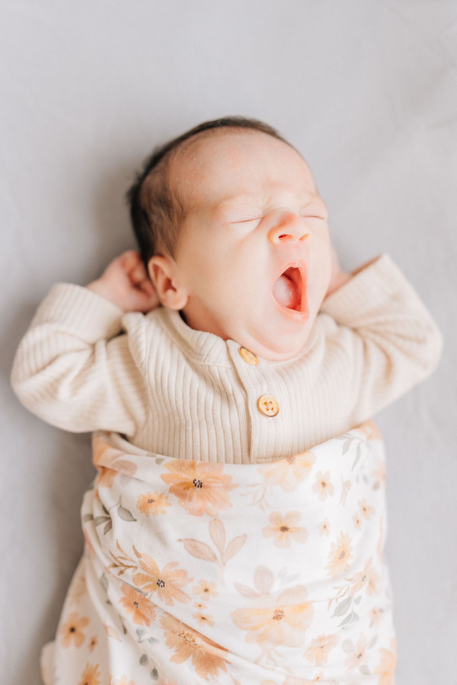 A sleepy newborn baby in a cream sweater and floral swaddle yawns while lying on a light background.